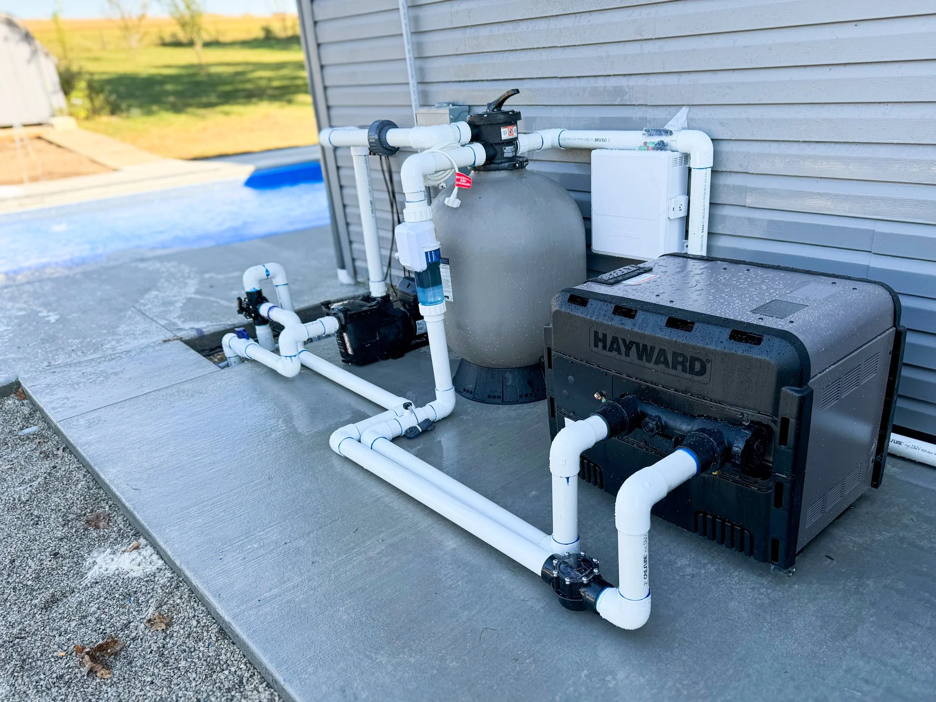 Pool equipment including a Hayward pool pump, filter, and filtration system with connected white PVC pipes on a concrete slab outside a house.