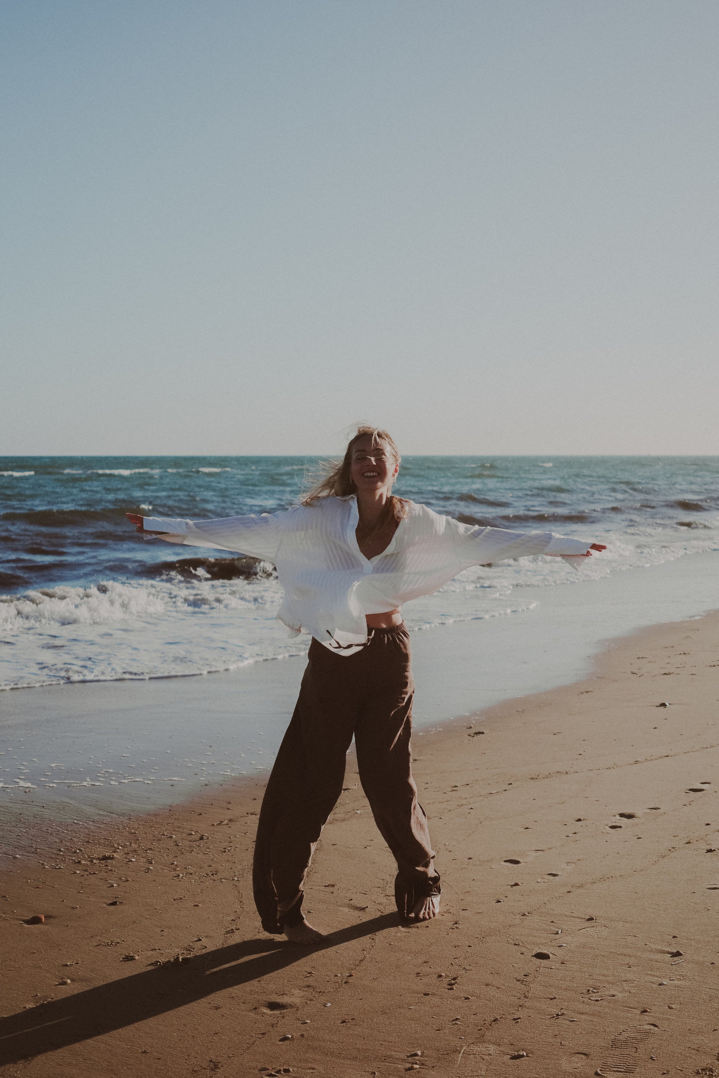 A woman standing on a sandy beach with arms outstretched, smiling, wearing a white blouse and brown pants, with ocean waves in the background and clear sky.