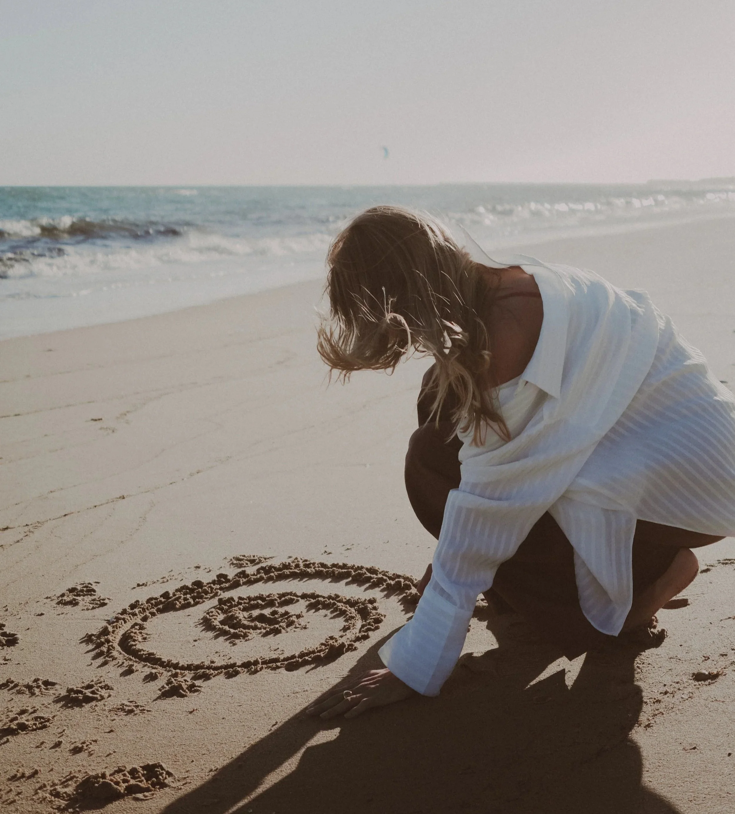 Person drawing a heart in the sand on the beach with the ocean in the background.