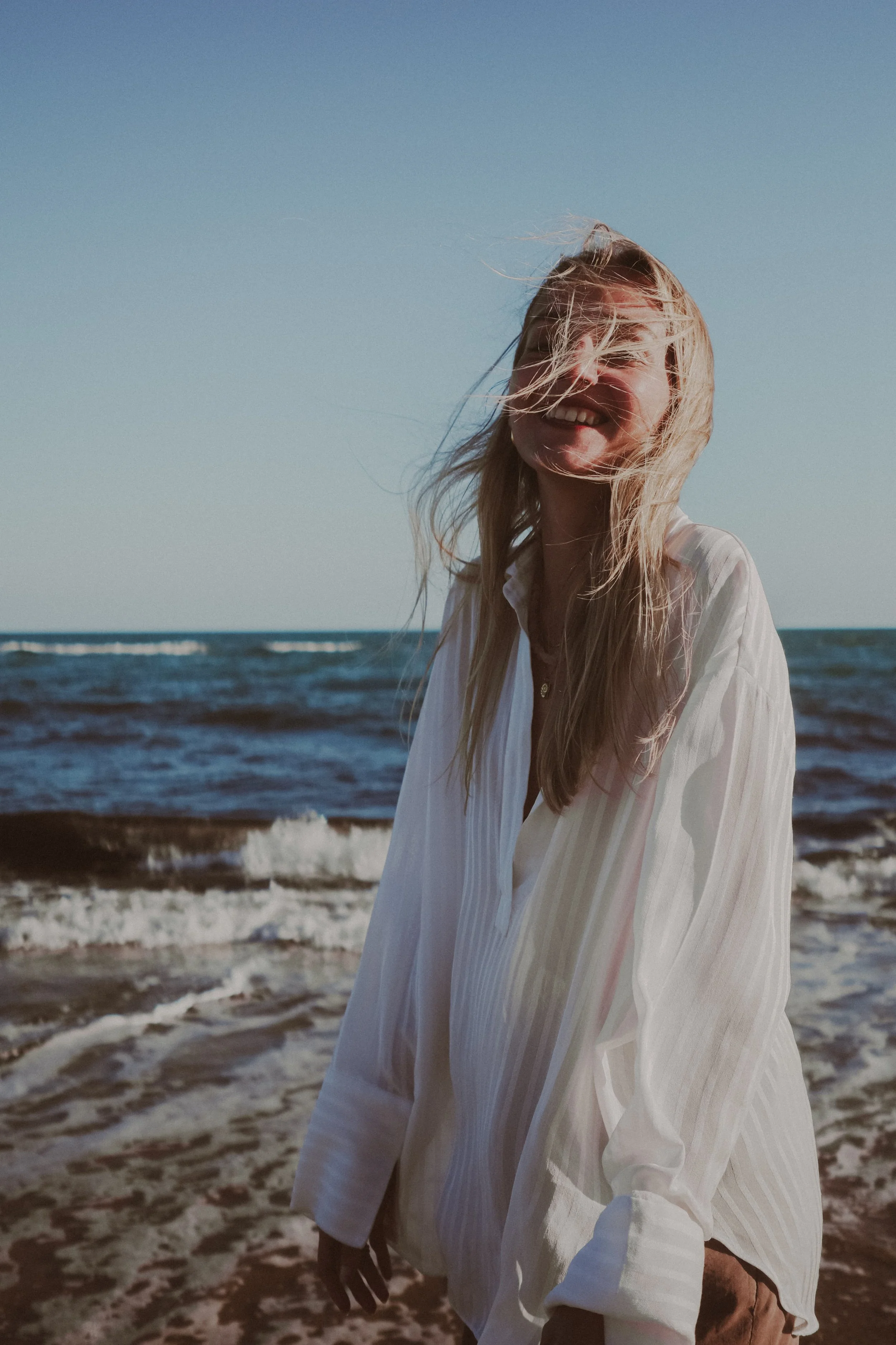 A smiling woman with blonde hair standing on a beach with ocean waves in the background, wearing a white shirt.