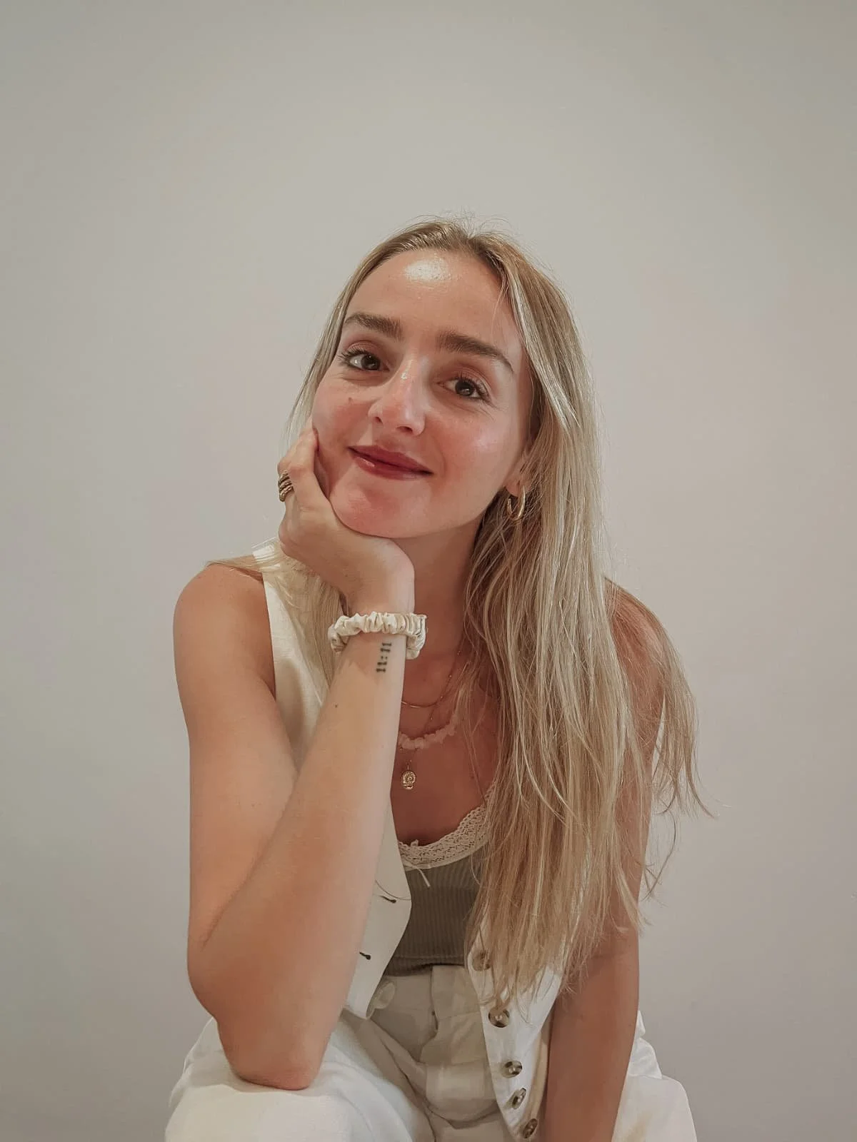 A woman with long blonde hair and fair skin leaning on her hand, smiling softly, wearing a beige top and white pants, set against a plain, light-colored background.