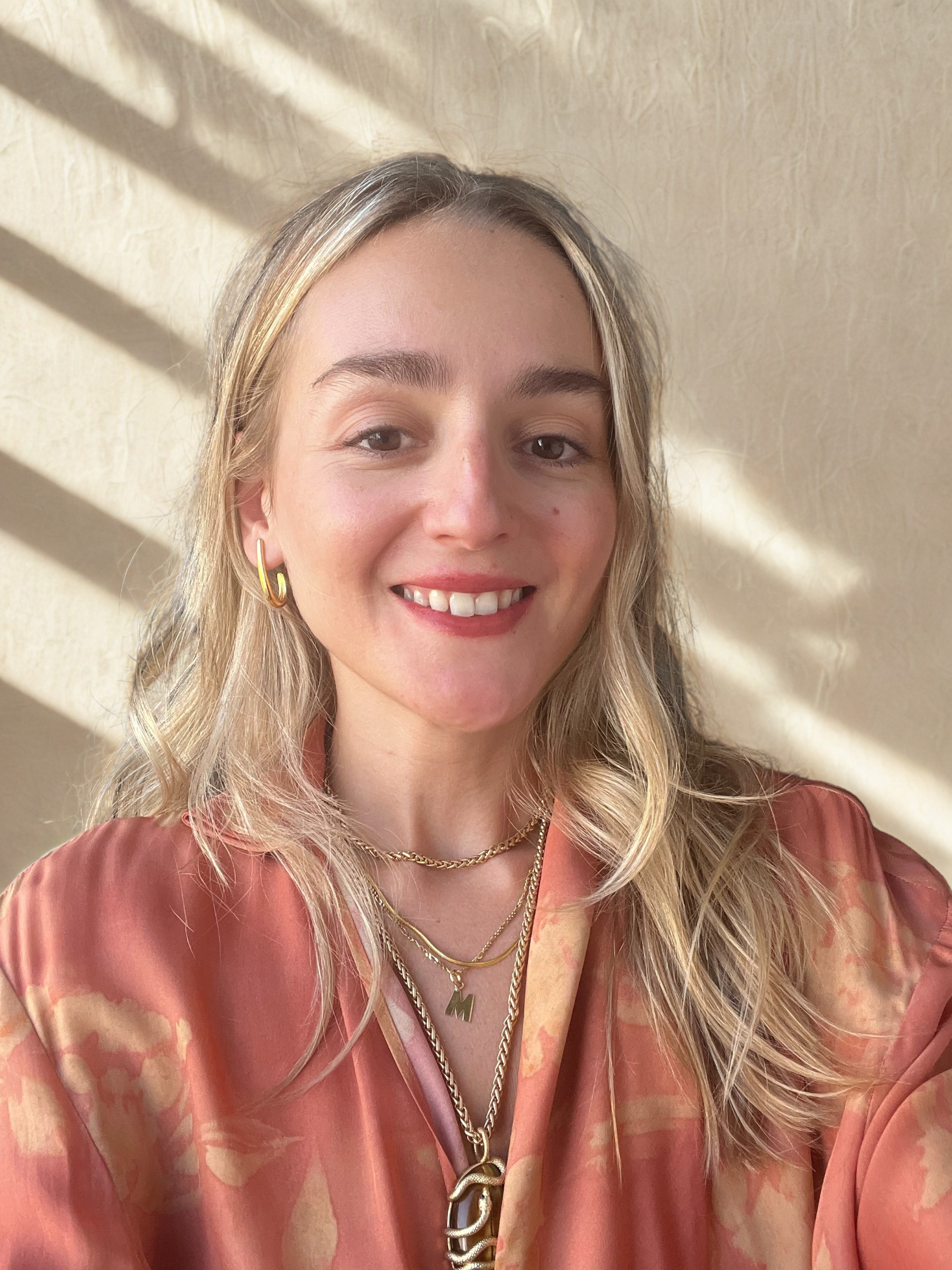 A young woman with wavy blonde hair, smiling, wearing gold hoop earrings, multiple layered gold necklaces, and a silky peach-colored blouse, with sunlight casting shadows on a beige wall behind her.