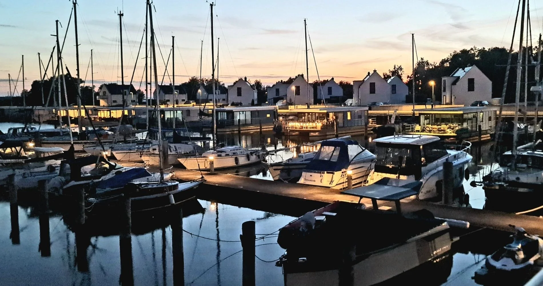 Abendstimmung im Yachthafen Perle: Die modernen Floating Houses spiegeln sich mit den Booten im ruhigen Hafenbecken. Warmes Licht aus den Häusern, Segelmasten und schwimmende Häuser direkt am Wasser – Urlaub auf dem Hausboot auf Usedom.