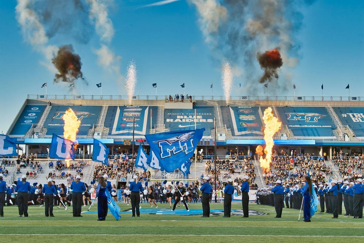 A football game at a stadium with players, cheerleaders, and spectators; blue flags, pyrotechnics, and smoke on the field and in the sky