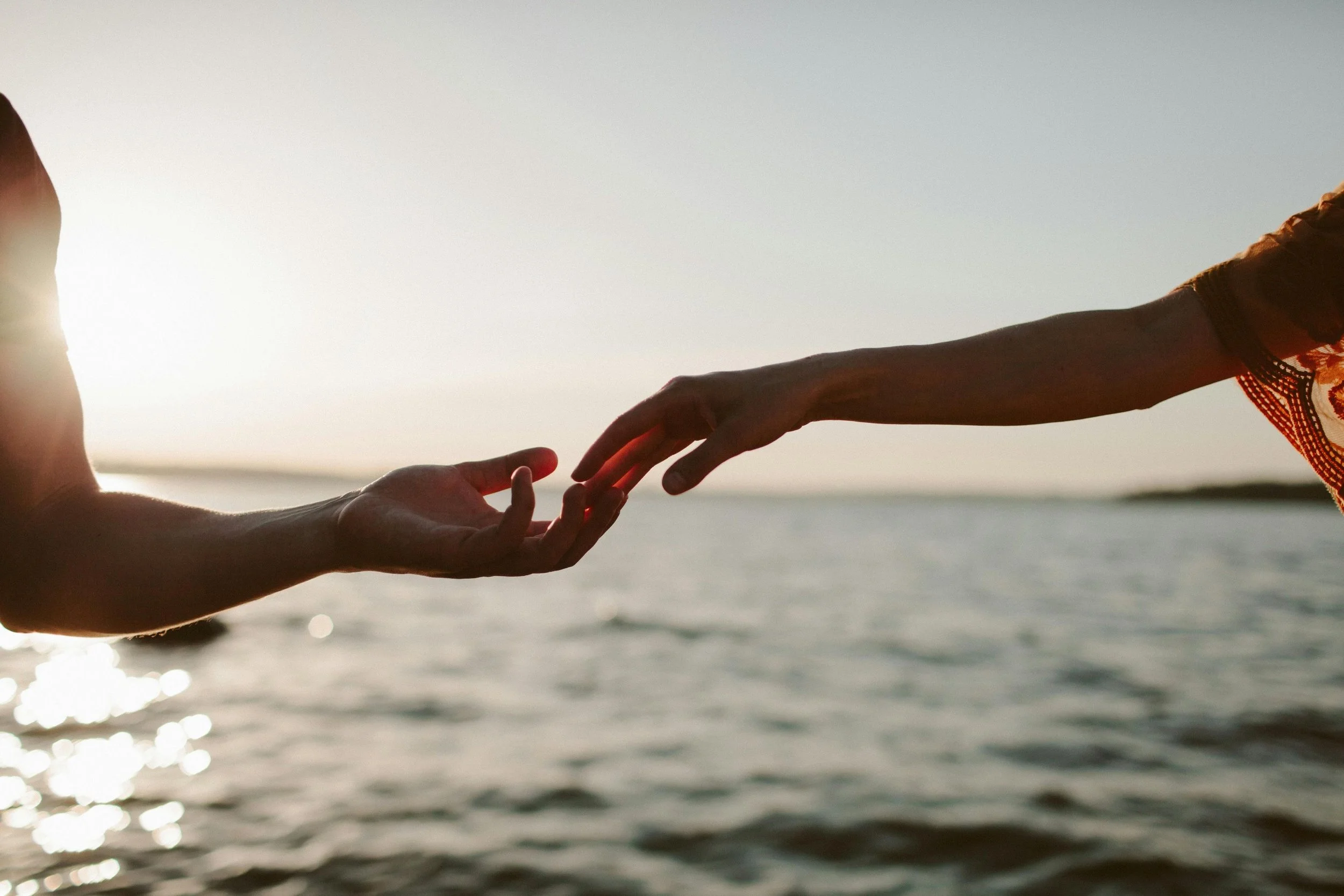 Two hands reaching toward each other by the water representing intuitive healing and human connection.