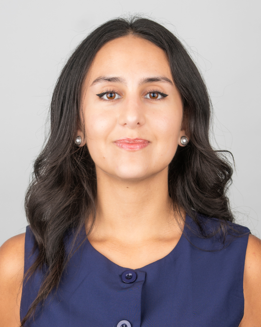 A woman with long dark hair, wearing a sleeveless navy blue top with buttons and pearl earrings, smiling softly against a plain light gray background.
