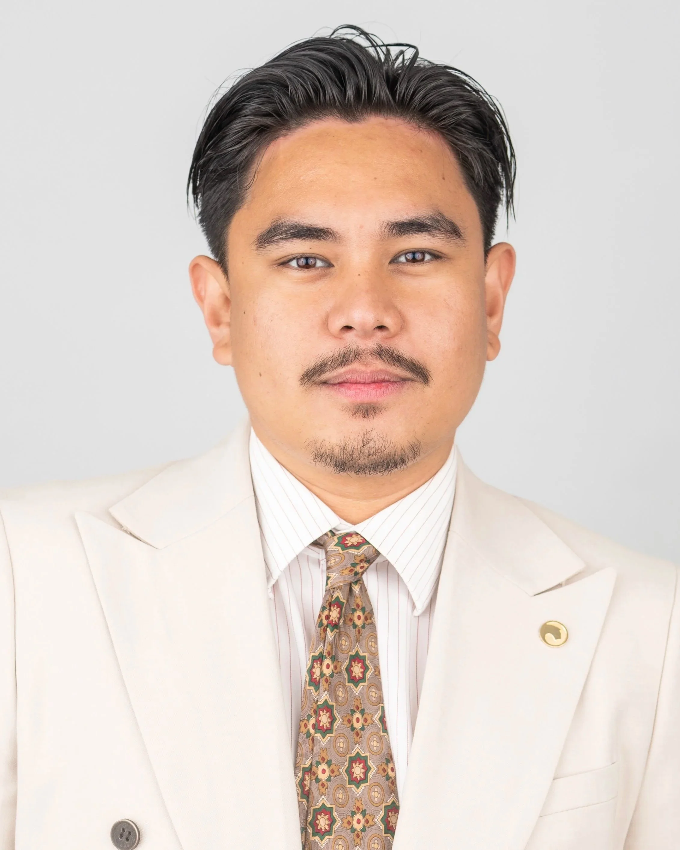 Close-up portrait of a man with white shirt, beige blazer, patterned tie, and dark hair styled back, standing against a plain light background.