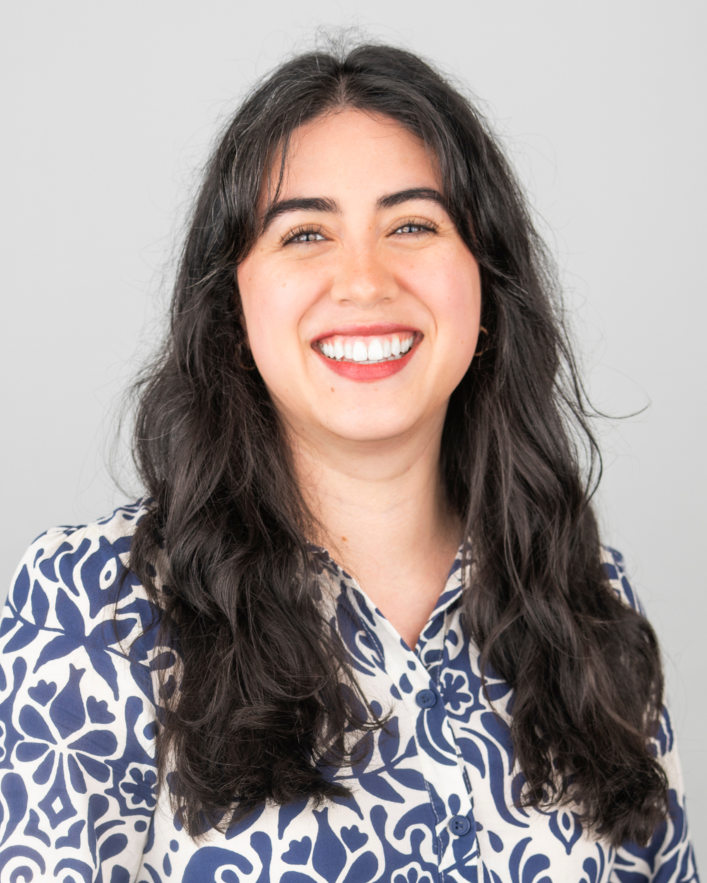 A woman with long dark wavy hair smiling, wearing a white and blue patterned blouse, against a plain light gray background.