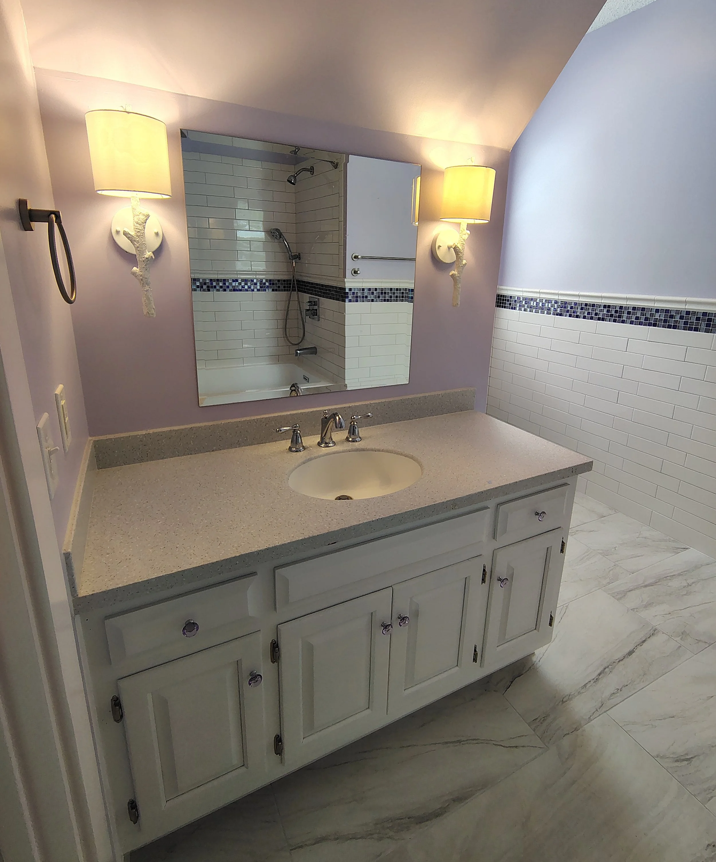 A beautifully renovated bathroom with a white vanity and countertop, oval sink, large mirror, two wall-mounted accent sconce lamps, purple walls, and a shower area with white tiles and a purple mosaic border.