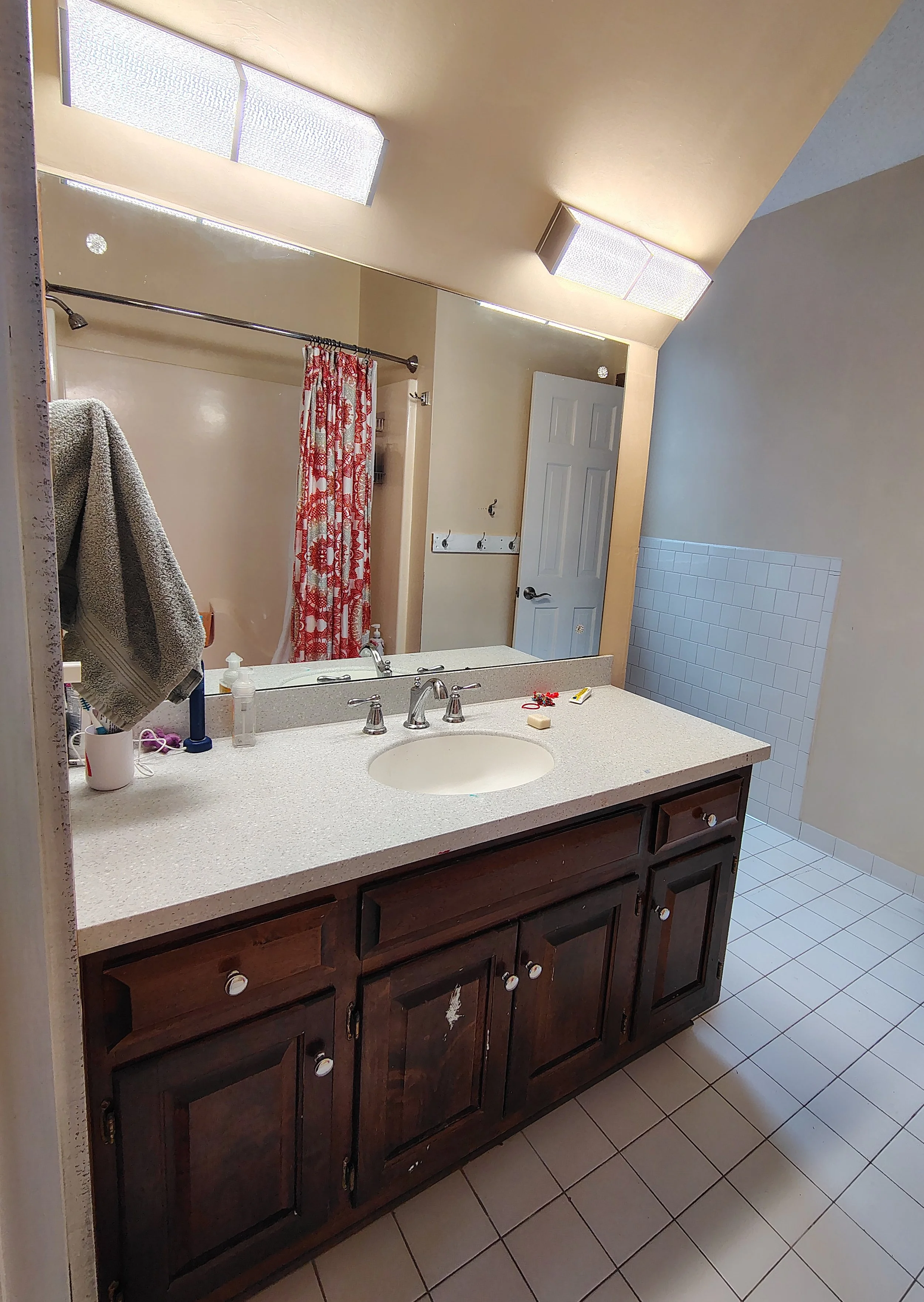 A bathroom with a wooden vanity, white countertop, and oval sink. Above it is a large mirror, with two ceiling lights reflected. To the left, a towel hangs on a hook, and in the background is a shower area with a red and white patterned shower curtain. The floor is tiled with square white tiles, and part of a wall with white tiles is visible.