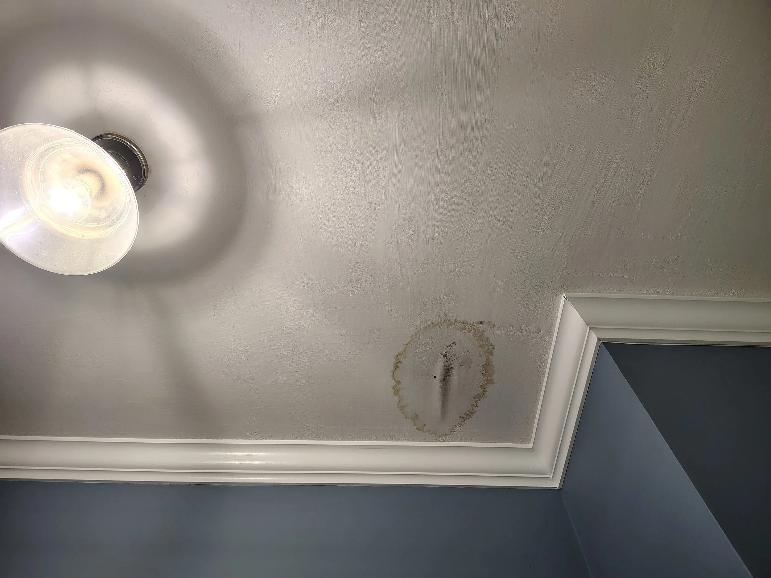 Ceiling with a light fixture and a water stain from leaking plumbing pipe.