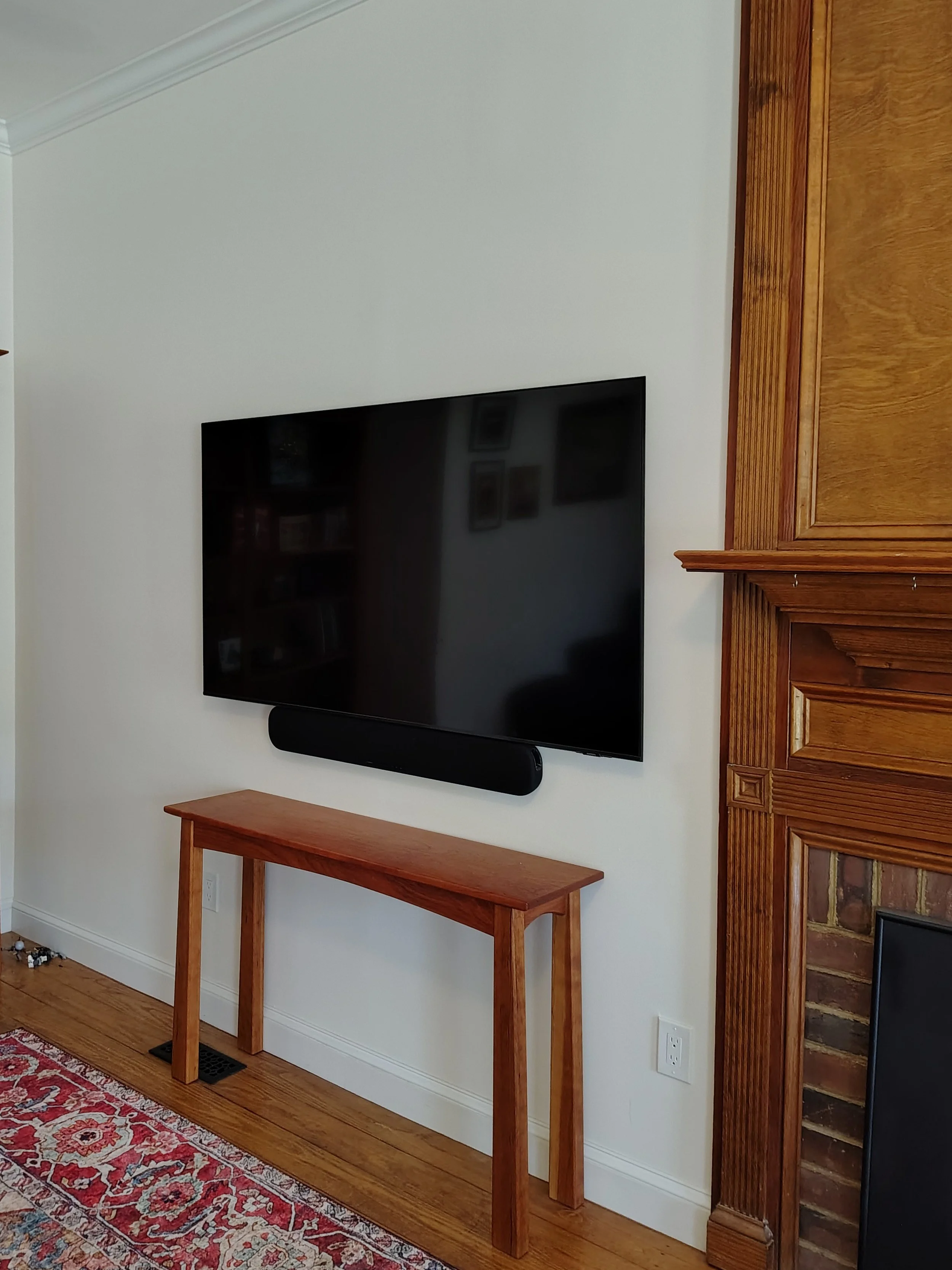 Renovated, rejuvenated and modernized living room corner with seamless drywall repair, a mounted flat-screen TV, soundbar below it, wooden console table, hardwood flooring, and a brick and wood fireplace.