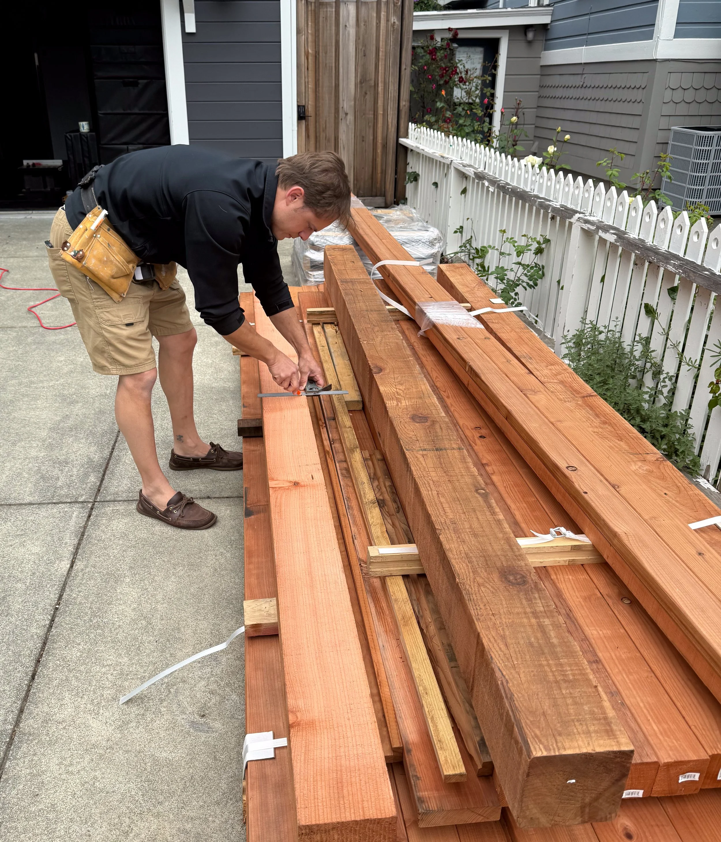 A man is working on a woodworking project outdoors, measuring and cutting pieces of wood on a large work surface, with a white picket fence and house in the background.