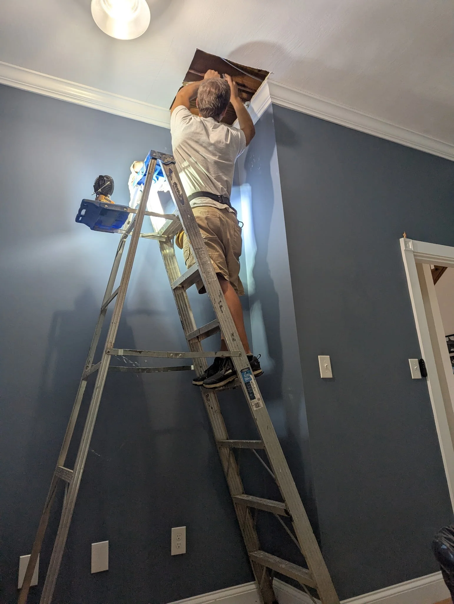 A person standing on a ladder installing or repairing ceiling drywall near a light fixture.