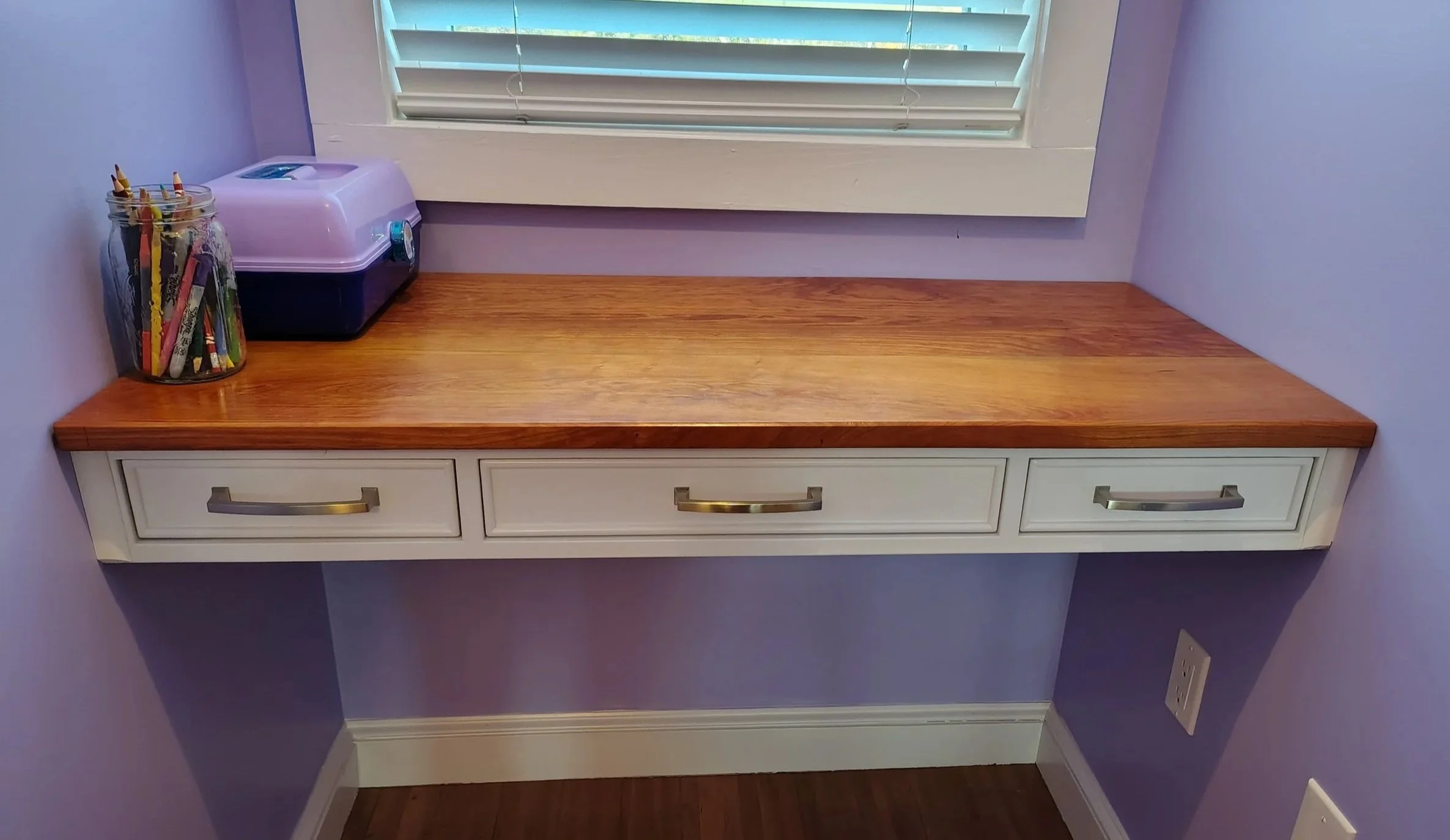 A custom built-in solid wooden desk with cherry wood top and three full-extension drawers beneath a window with white blinds, purple walls, a jar of colored pencils, and a small purple crafts box on the left side.