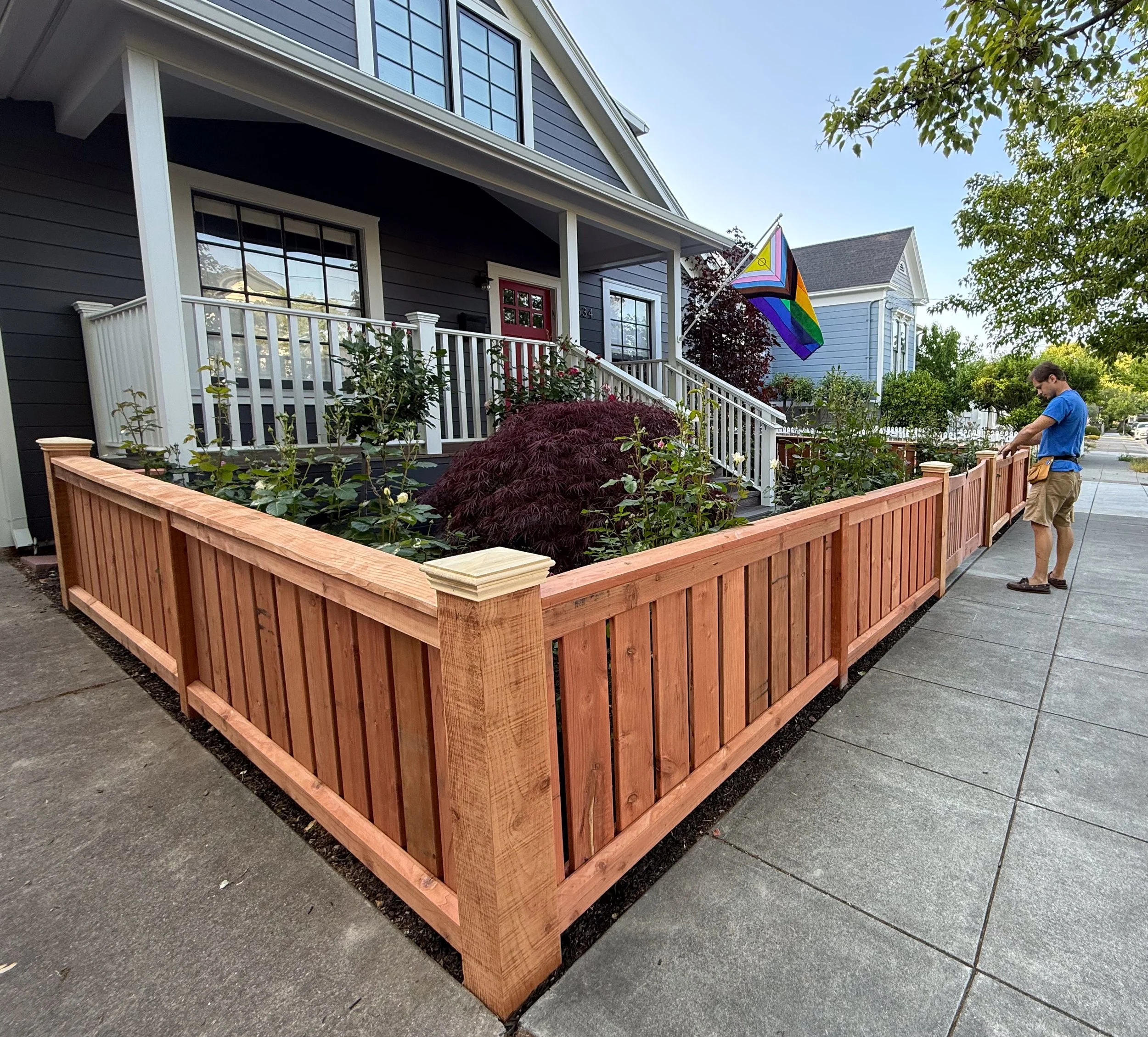 A house with a small garden enclosed by a newly installed craftsman style redwood wooden fence. The house has a front porch with white railings, a red door, and a rainbow pride flag flying nearby. There are trees and other houses in the background.