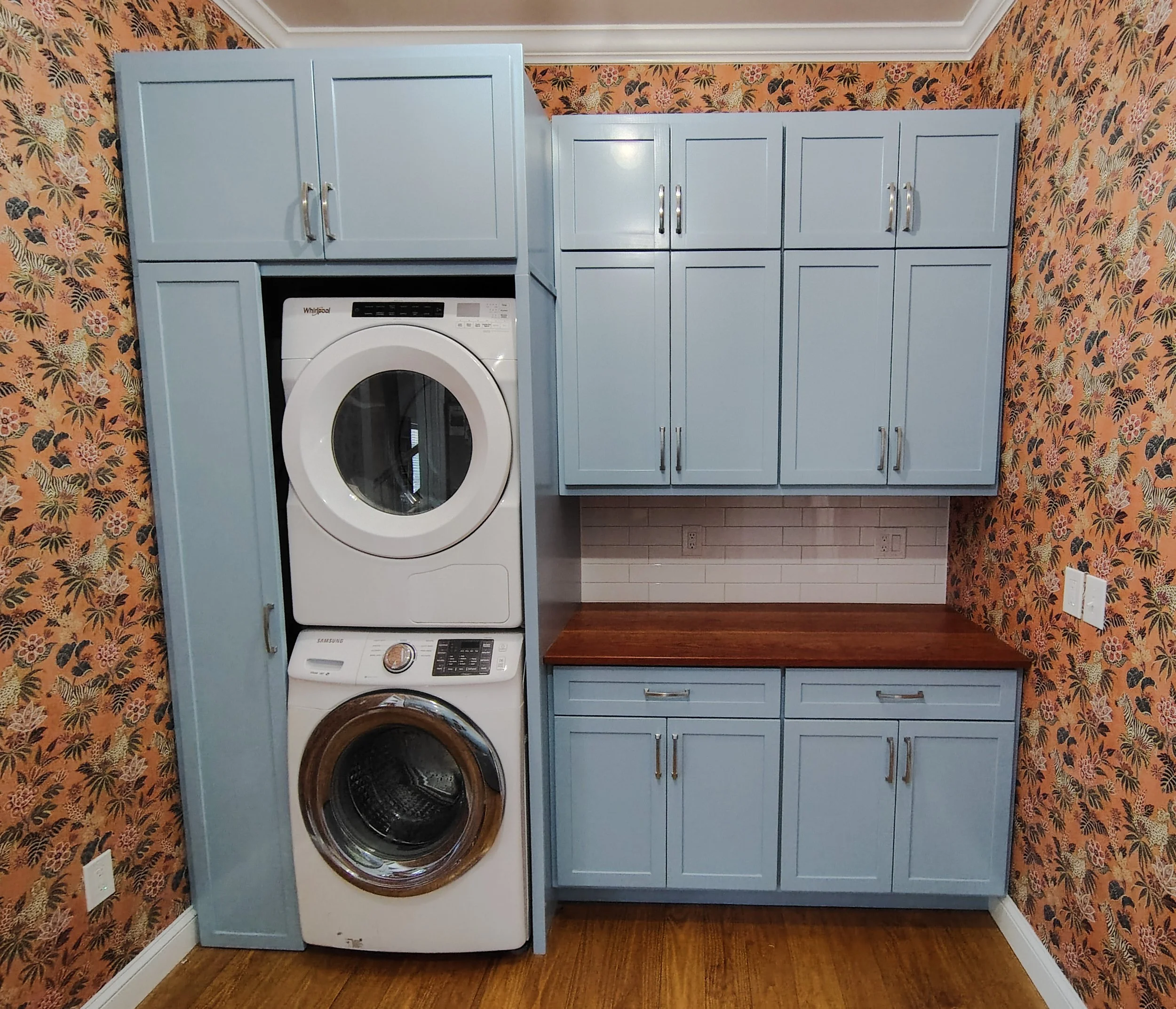 Custom solid wood cabinets in a laundry room with solid cherry wood counter tops and trendy wallpaper.  It has a stacked washer and dryer, blue cabinets, wooden countertop, floral wallpaper, and electrical outlets.