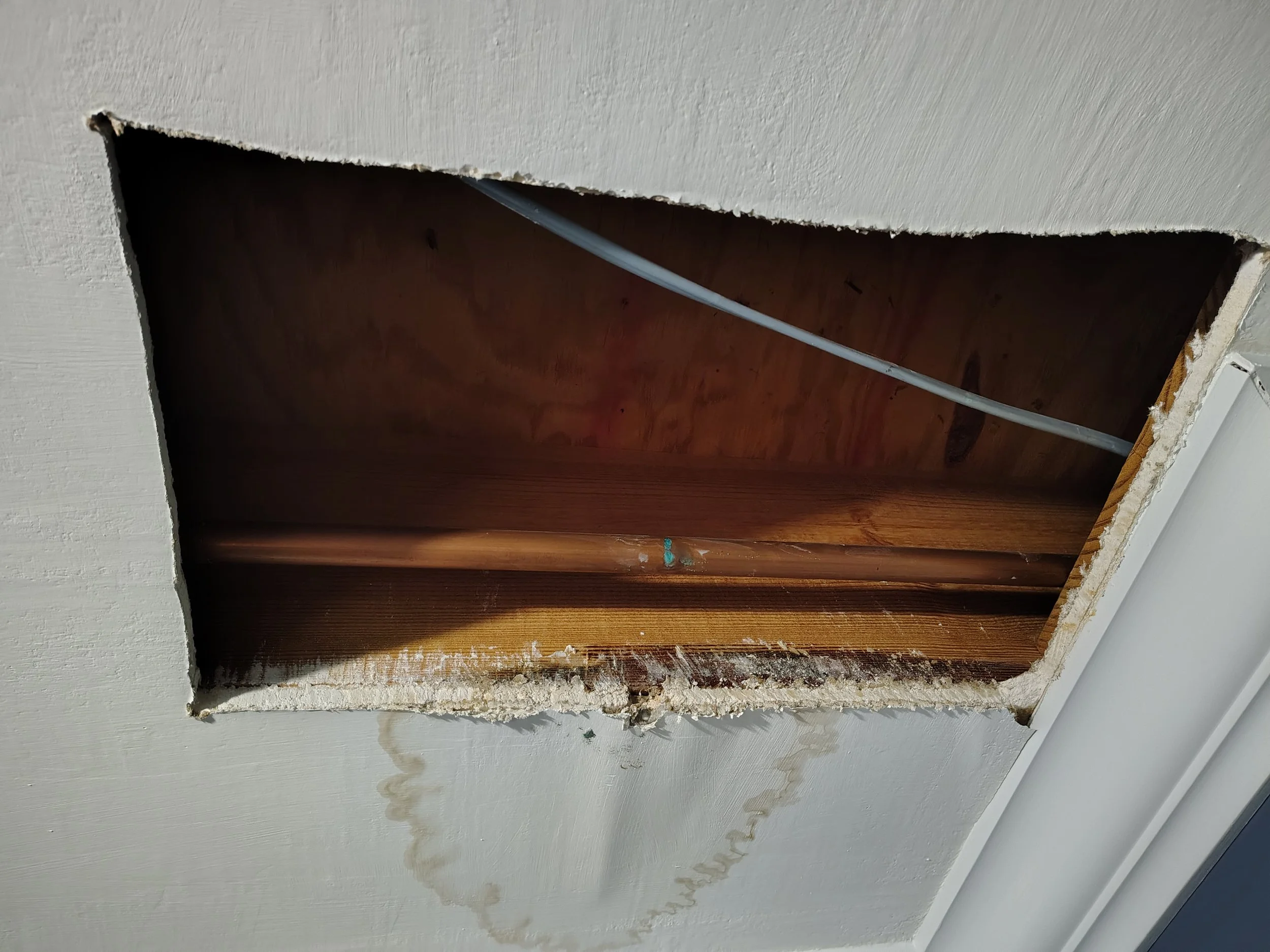 Ceiling damage from leaking water plumbing pipe with ceiling opening showing wooden beams and electrical wiring and leaking pipe inside a room.