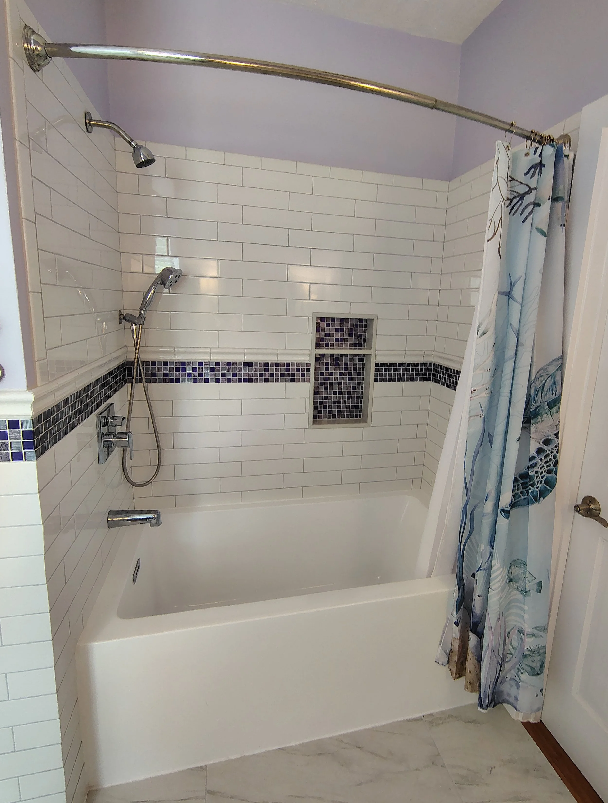 A beautifully renovated bathroom with a white bathtub, white subway tile walls with black and purple mosaic accents, a showerhead, a handheld shower, a shower curtain with a marine theme, and a wall niche.