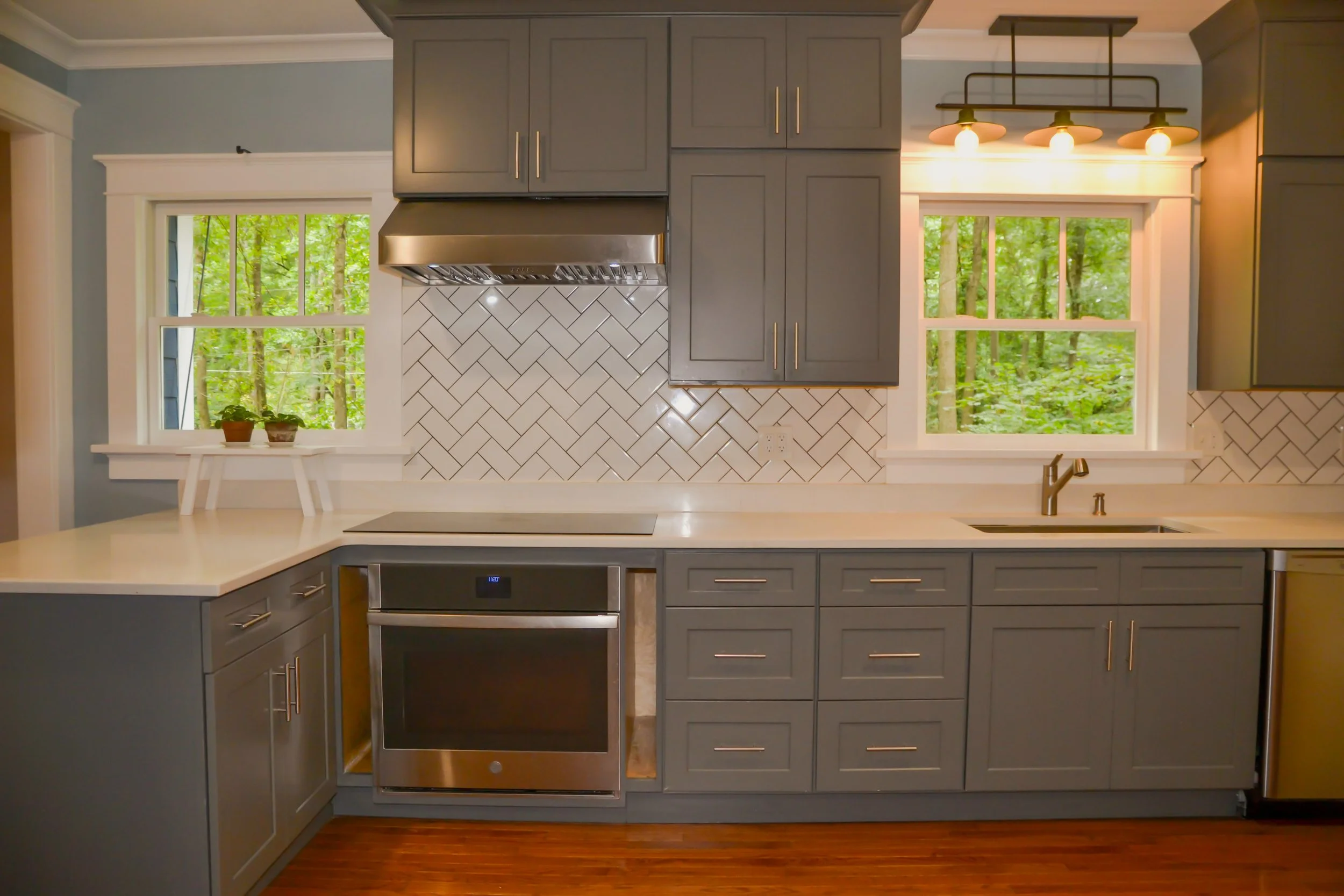 Custom modern renovated kitchen with gray cabinets, white countertops, a stainless steel oven, and a herringbone white tile backsplash. There are two windows with green trees outside and a wooden floor.