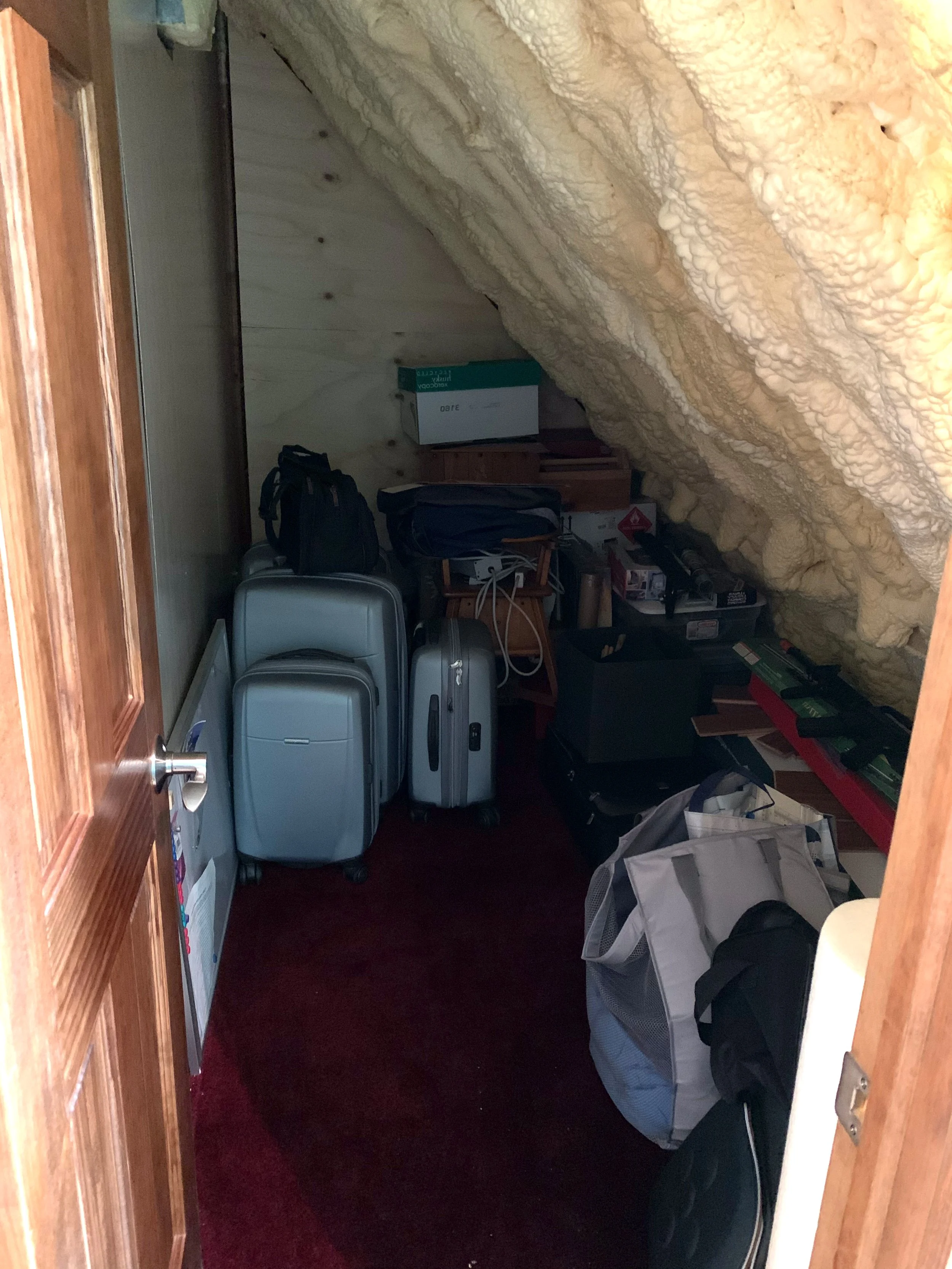 Cluttered storage room with suitcases, bags, boxes, and miscellaneous items on carpeted floor and shelves.