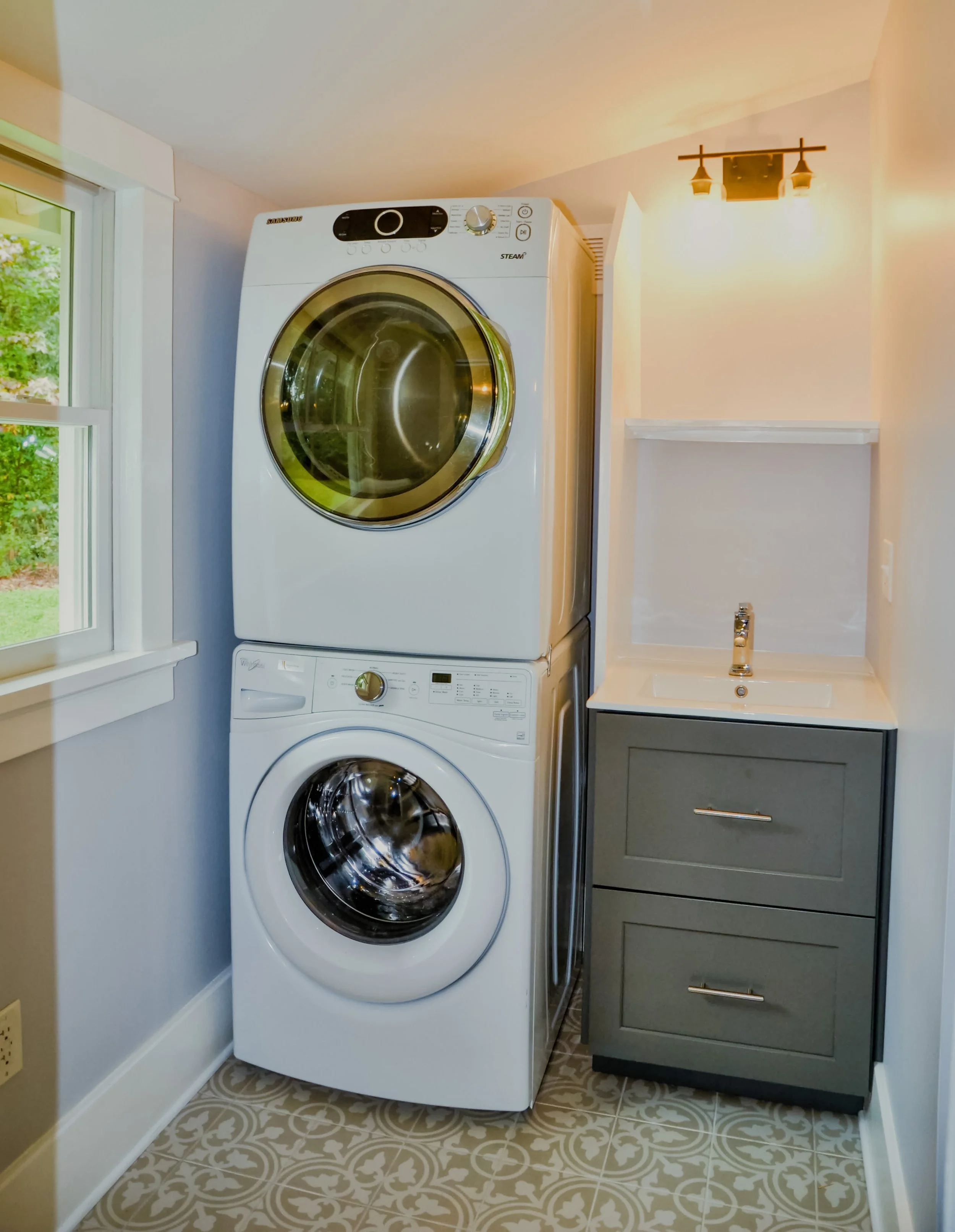A renovated modern custom laundry room with a stacked washer and dryer, a small sink with a cabinet, a window, wall-mounted light fixtures and trendy tile floor.