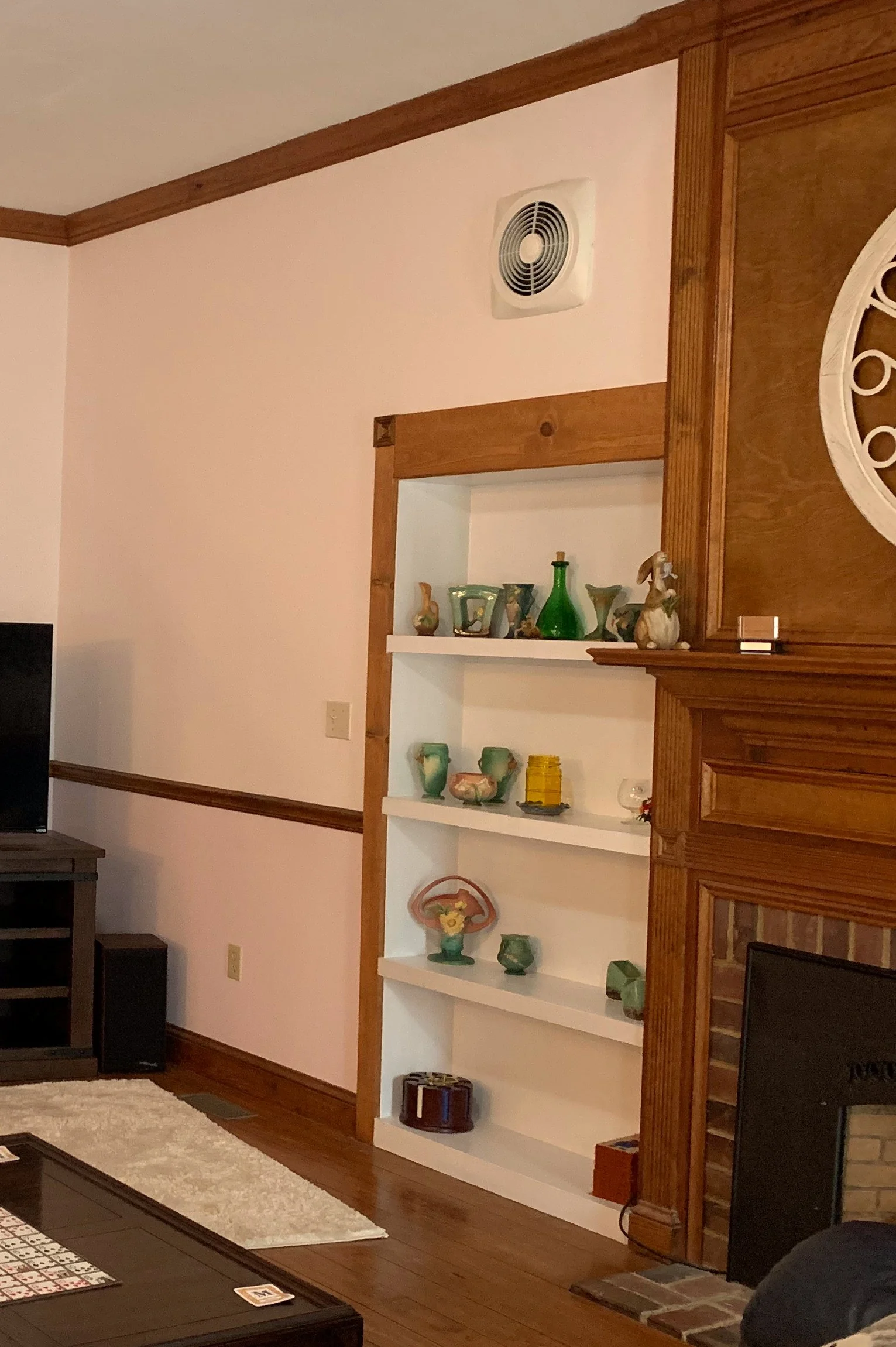 Dated looking living room before renovations with built-in wooden shelves containing decorative glassware and pottery, a wood-paneled fireplace, a TV on a stand, and a light-colored area rug.