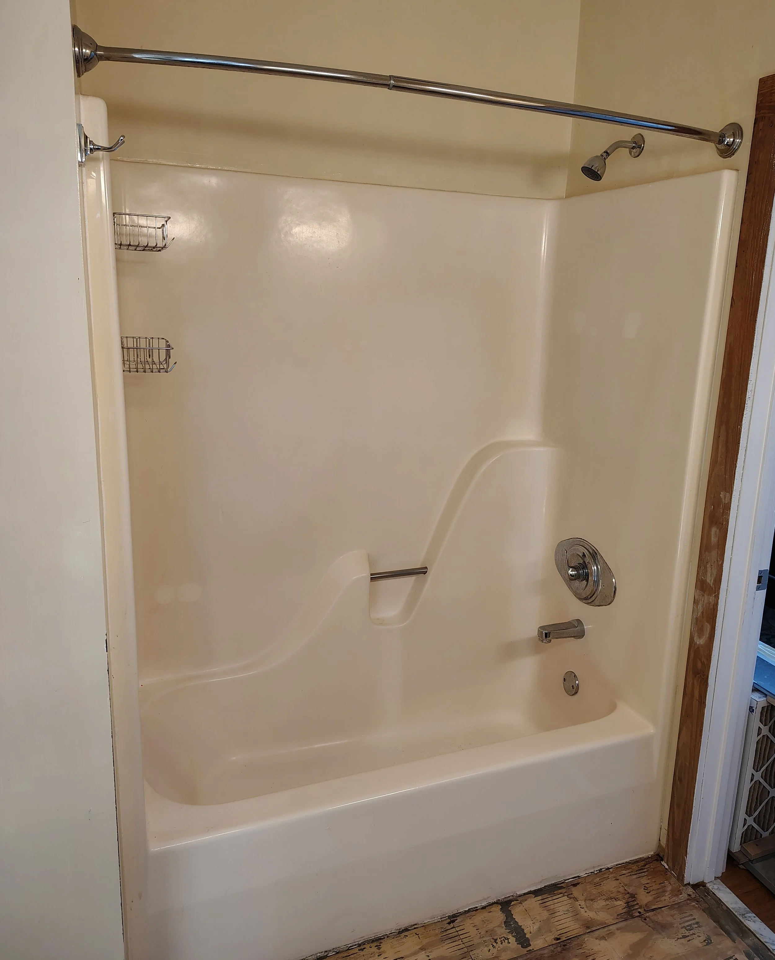Empty beige bathtub shower with a chrome showerhead, faucet, and two small wire shelves on the wall, surrounded by a beige wall and wooden floor.