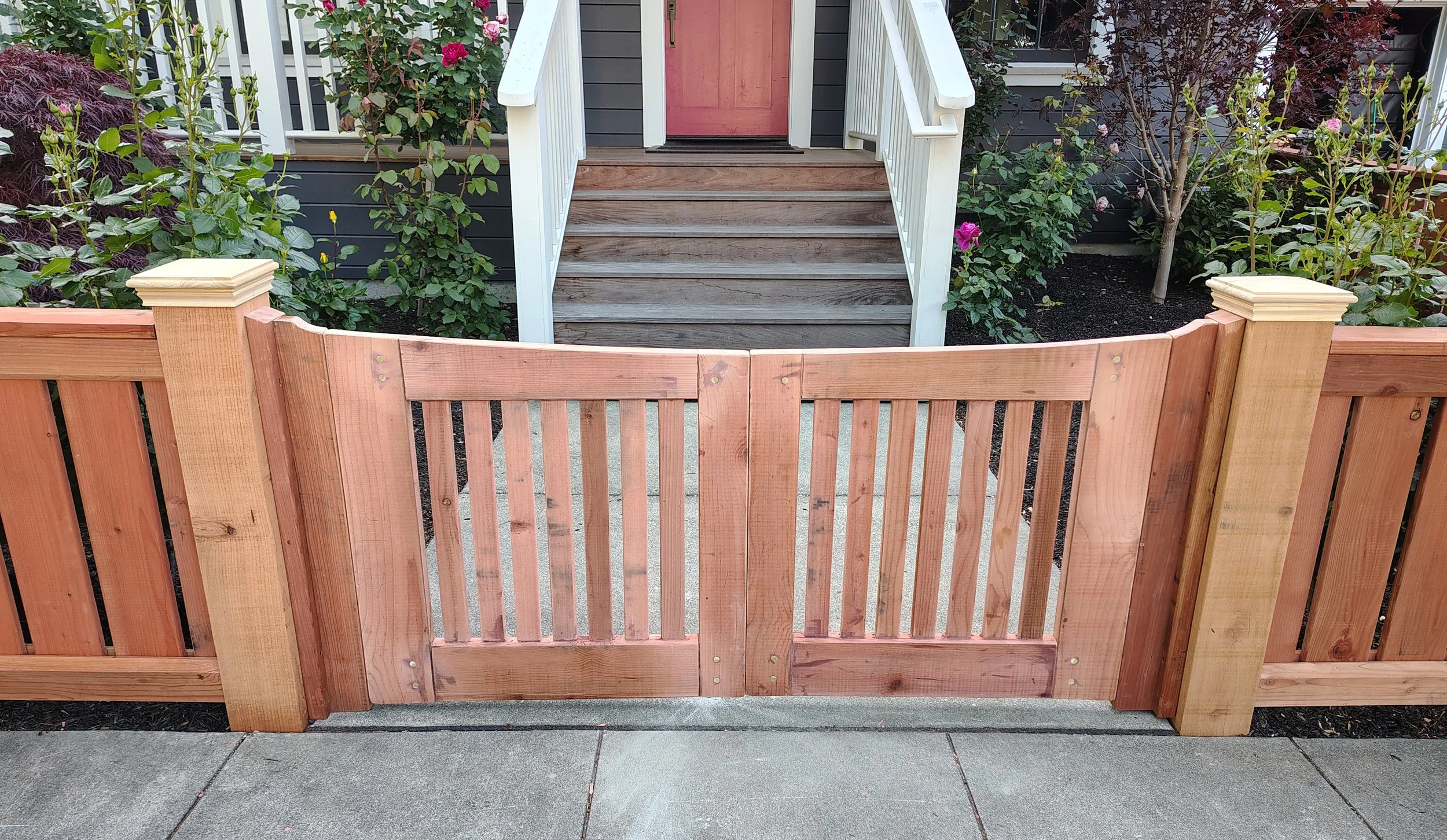 A solid wood craftsman style redwood wooden gate installed on a new craftsman fence at the sidewalk leading to the stairs of a house, with plants and flowers on either side of the stairs.