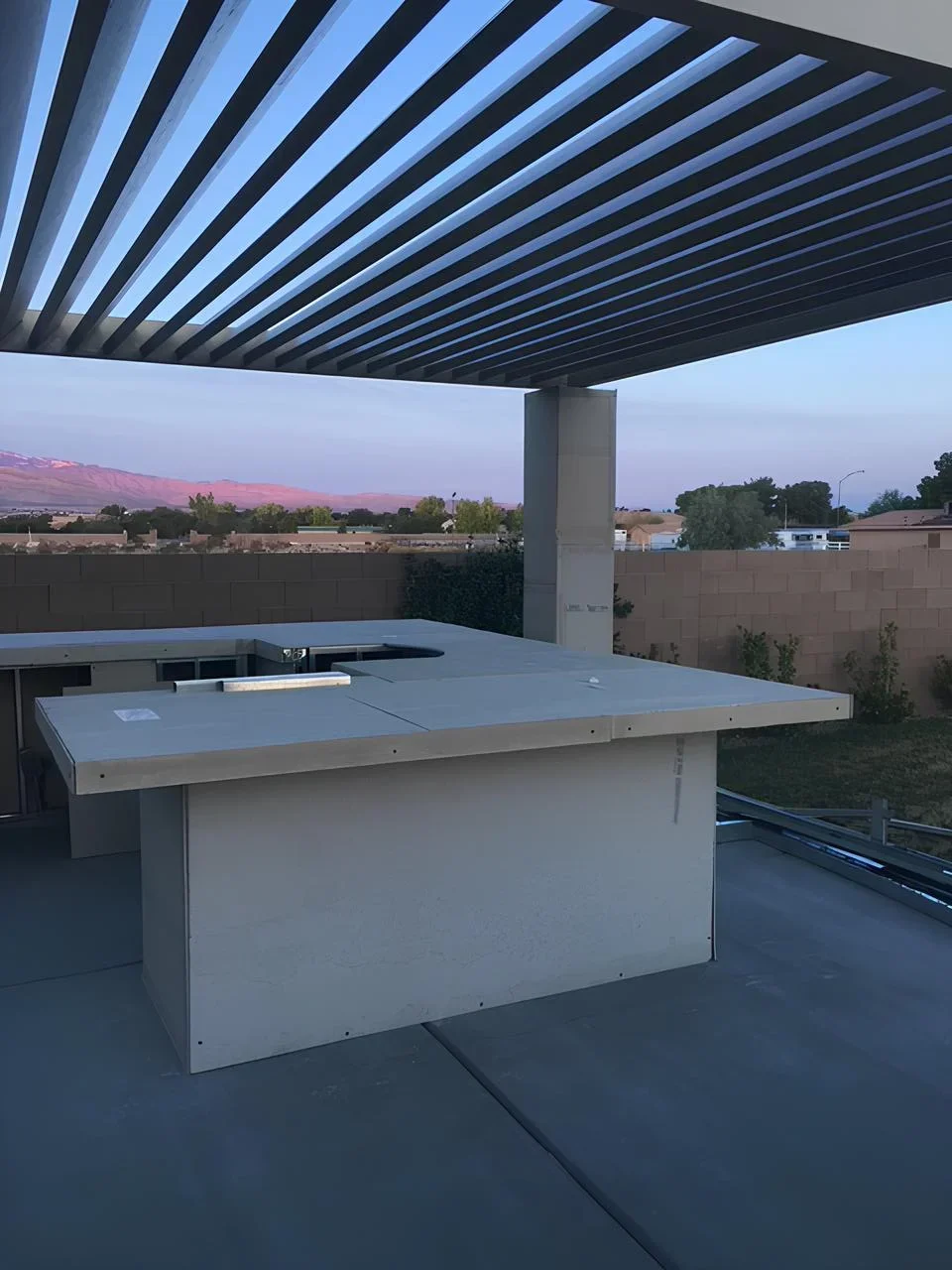 Empty outdoor kitchen area with a concrete countertop and a small sink, under a metal pergola, overlooking a suburban neighborhood with mountains in the distance.