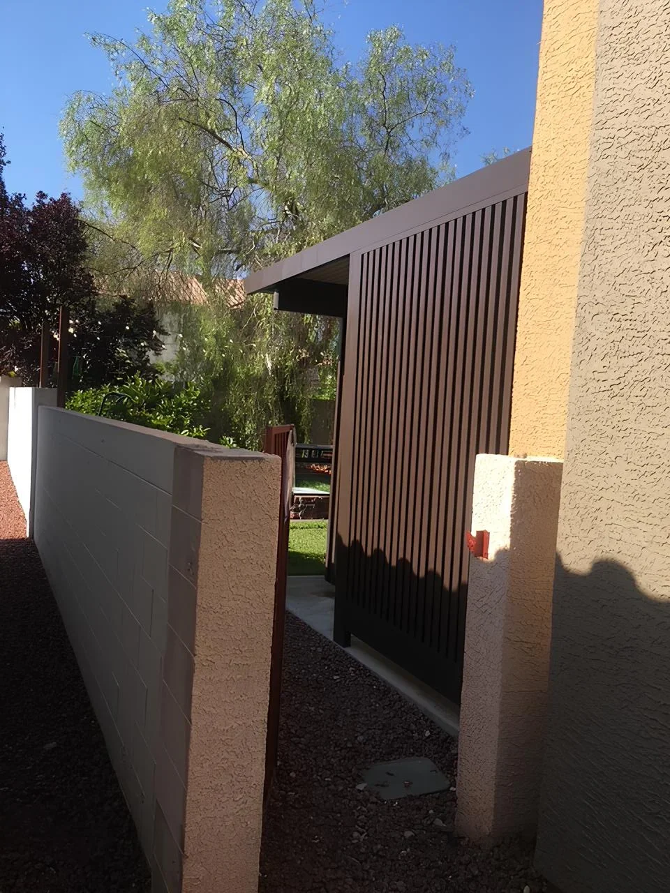 A modern outdoor backyard gate with brown slatted panels, beige stucco walls, a white concrete fence, and green trees in the background under a clear blue sky.