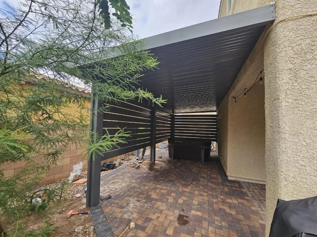 Covered patio area with brick paving, a black slat privacy wall, string lights, and a beige textured exterior wall of a house, with some construction tools and equipment present.