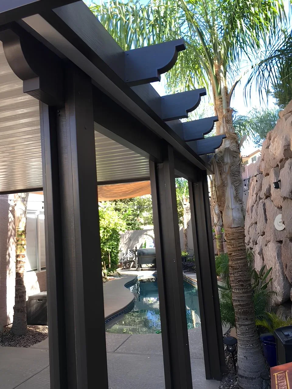 Part of a modern backyard with a pool, palm trees, and outdoor seating, seen through a black metal structure with a decorative top edge.