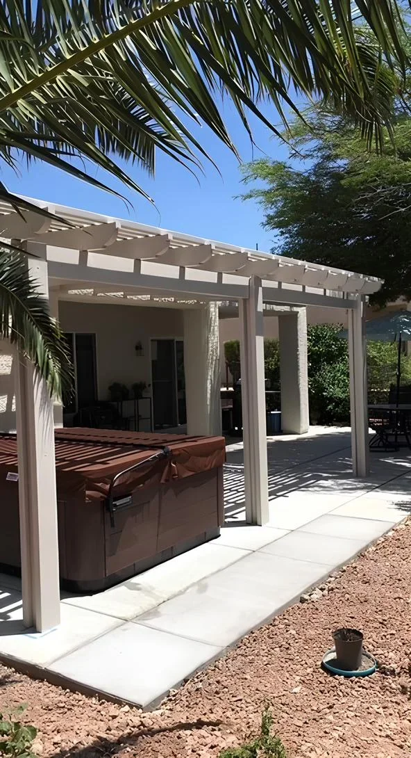 Patio with a hot tub under a pergola, surrounded by a house and trees, on a sunny day.