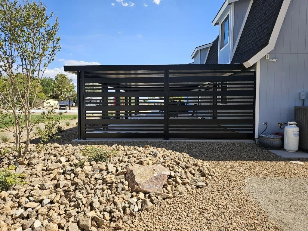 A modern house with a black horizontal slatted privacy fence on a gravel yard, next to a tree and some landscaping rocks, with a propane tank and tire nearby.