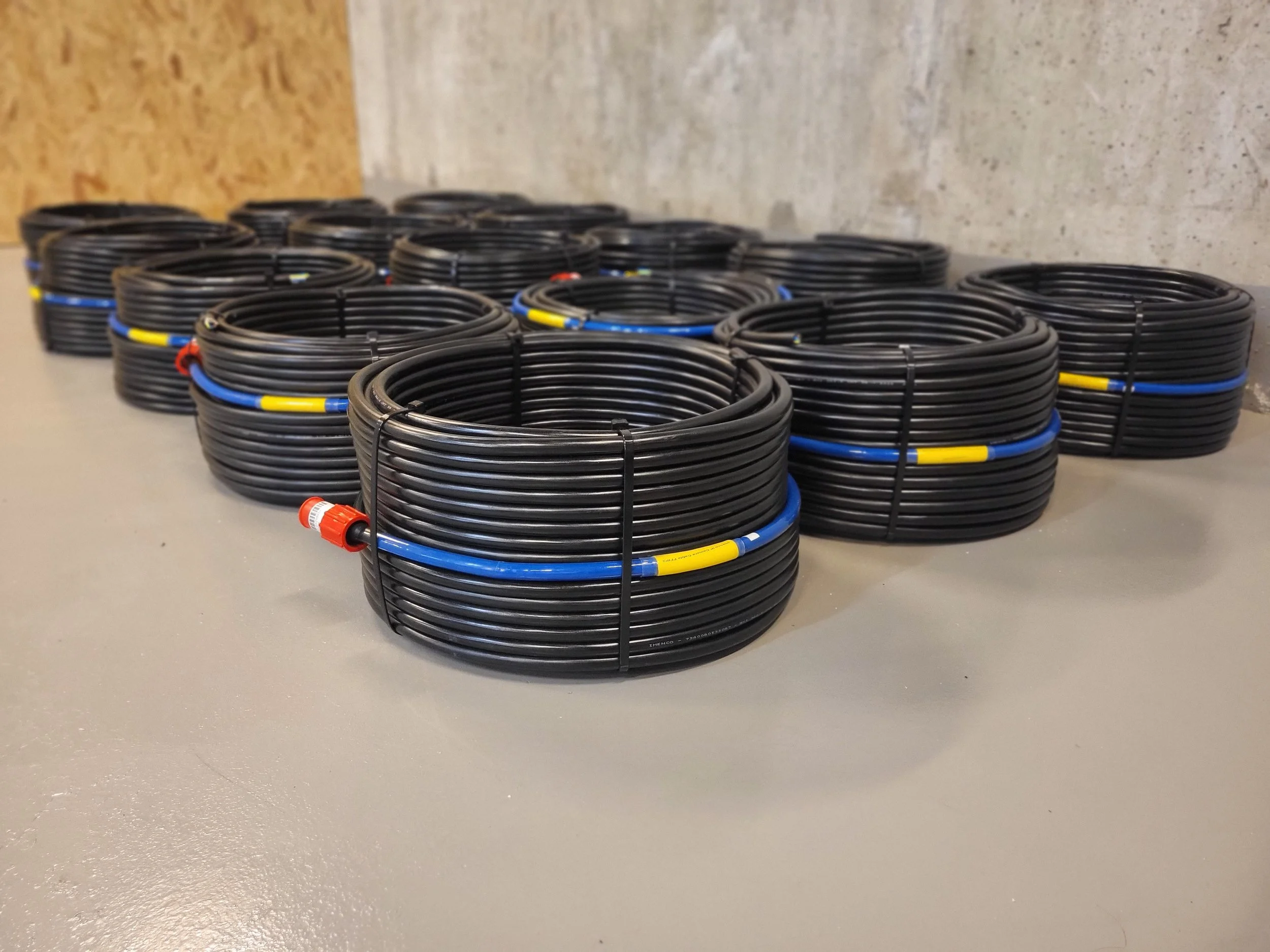 Multiple coiled black electrical cables with blue, yellow, and red colored connectors, neatly stacked on a gray floor against a background of unfinished wall. Subsea cables, fish farm
