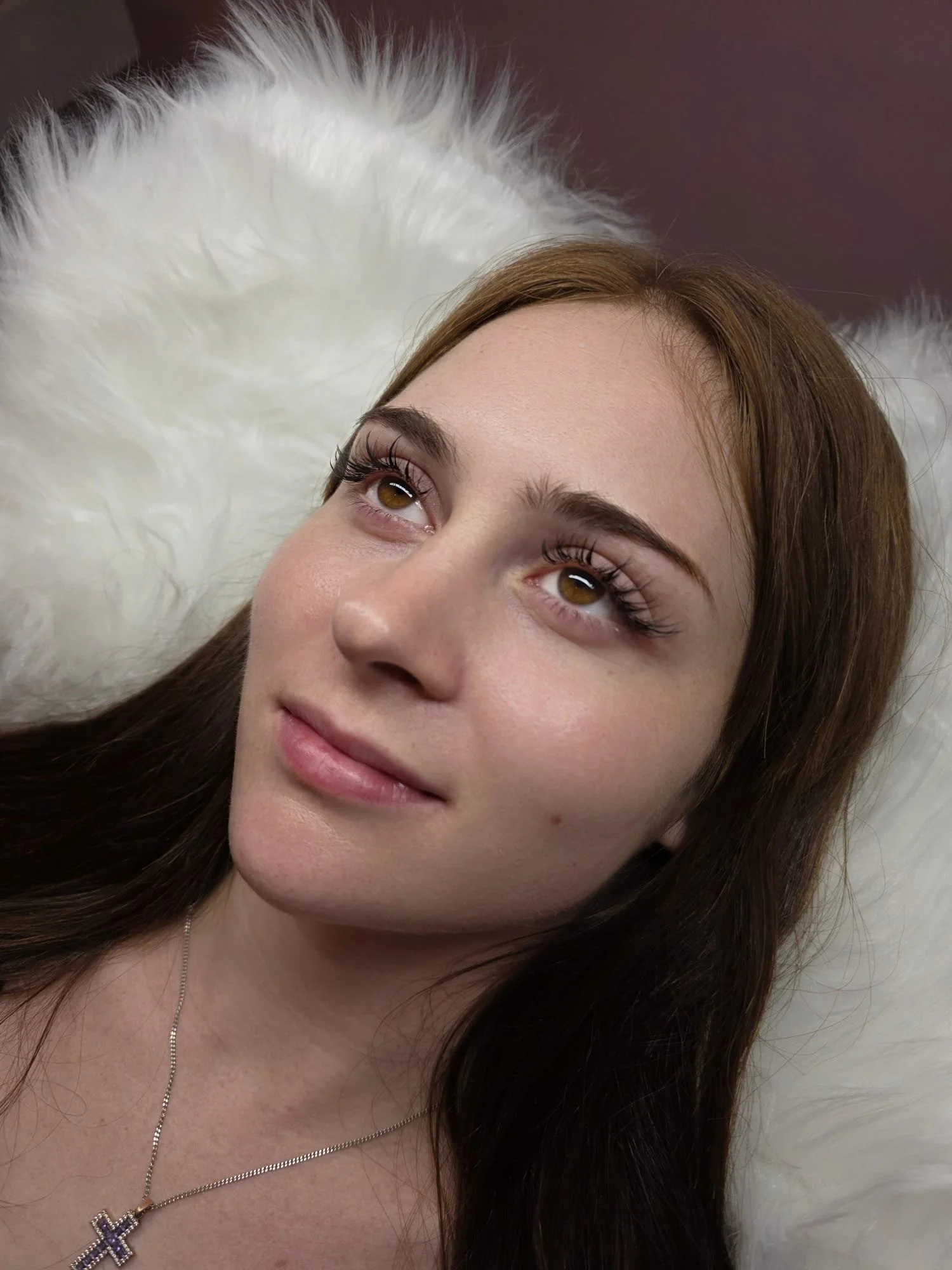 A young woman with long brown hair and wispy eyelash extensions lying on a white fluffy surface, looking up with a slight smile.
