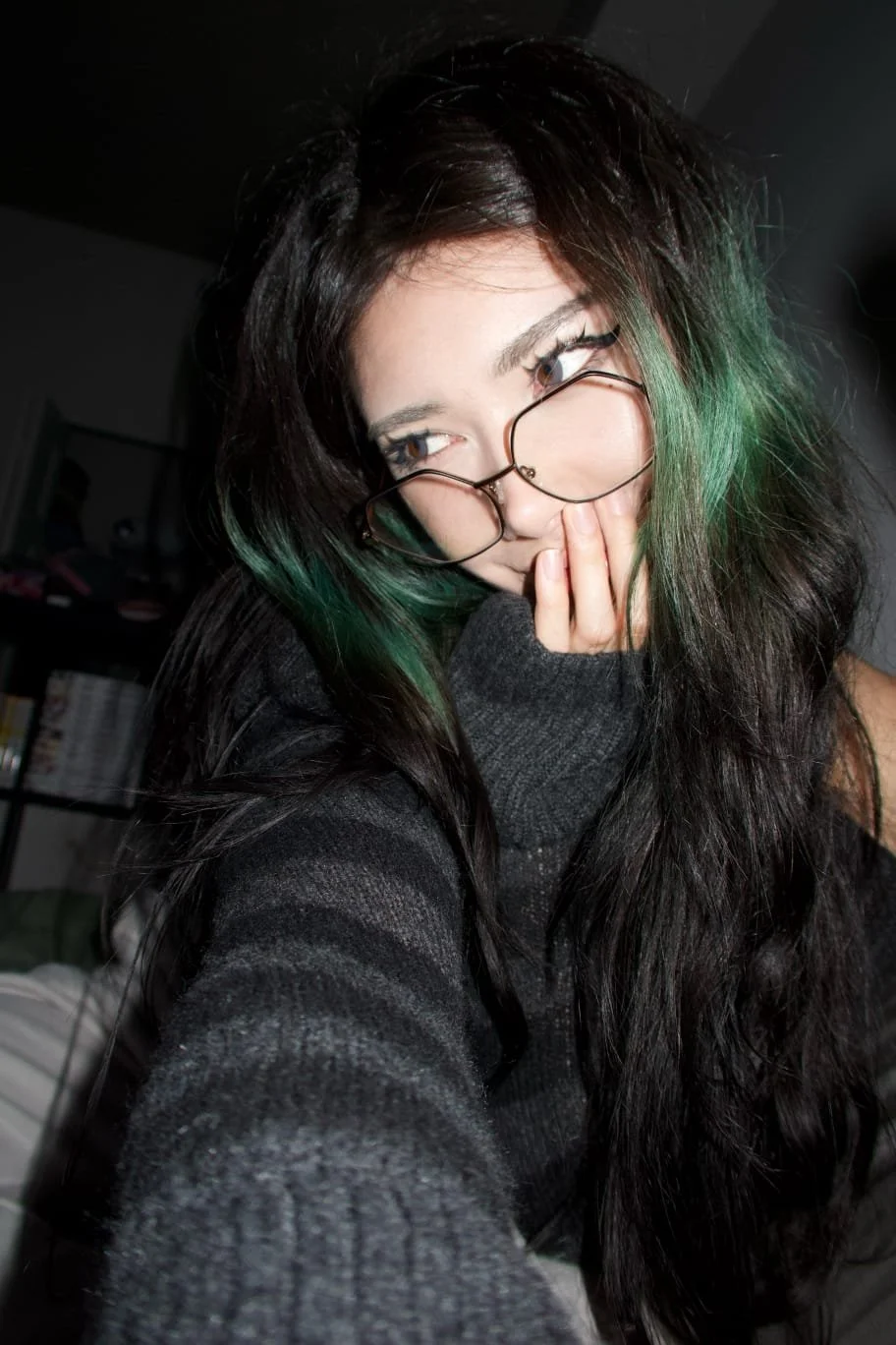 A young woman with black hair and green highlights wearing glasses and a black sweater, resting her face on her hand.