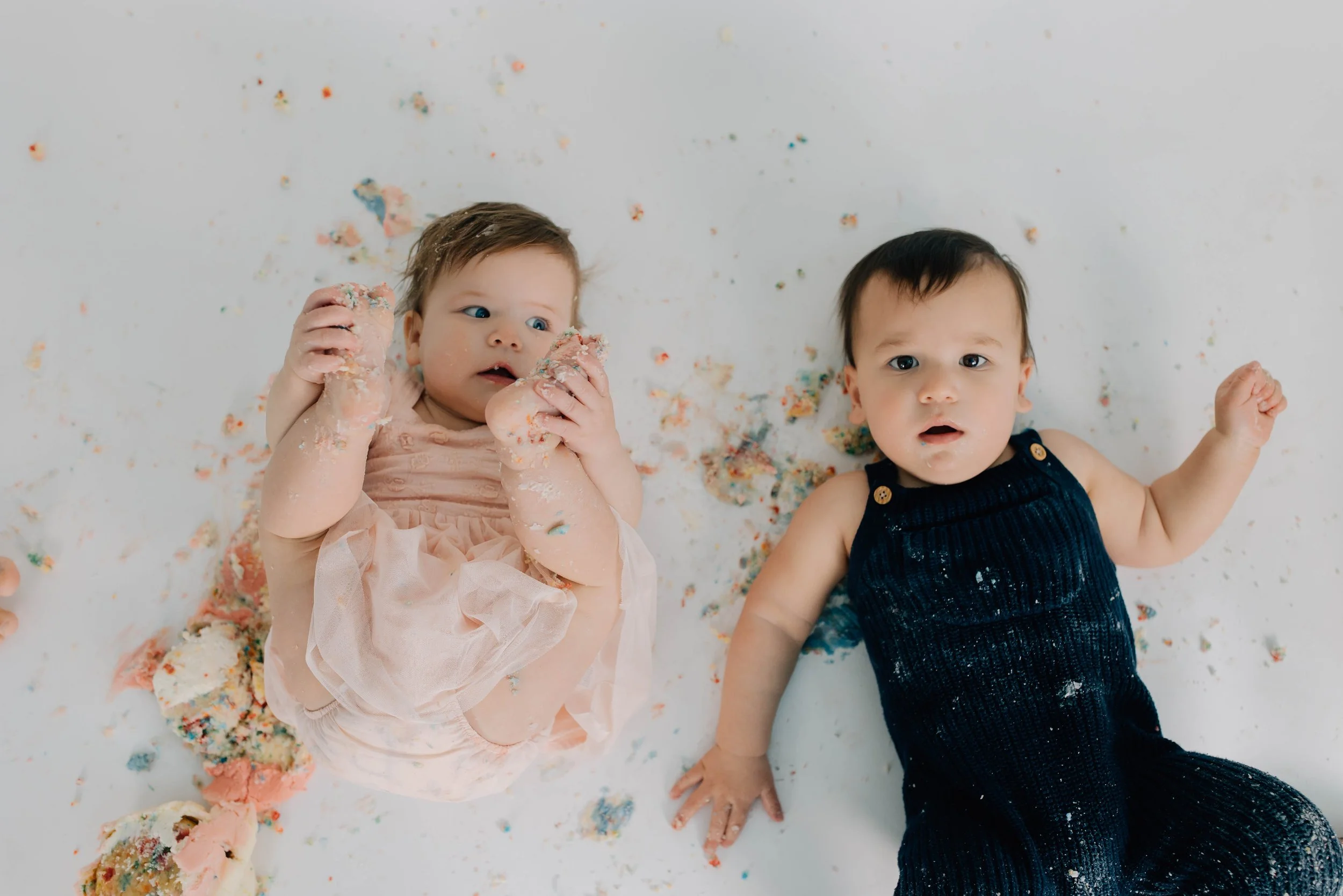 Two babies lying on a white surface covered in colorful cake and crumbs, one in a light-colored dress holding cake, the other in a dark outfit with cake on their clothes, with surprised expressions.