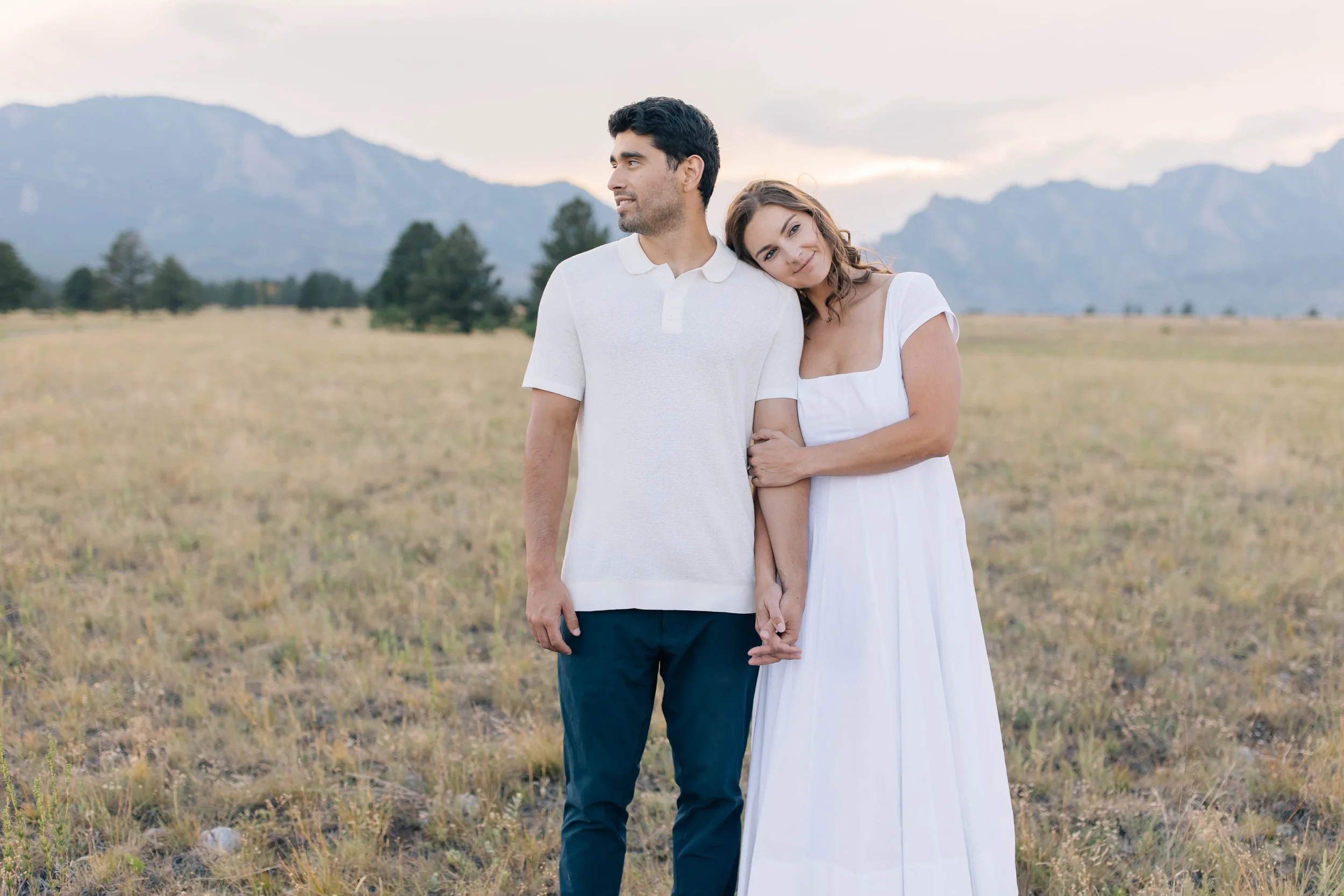 A couple standing in a field with mountains in the background, holding hands and leaning on each other, dressed in white and blue, in a serene outdoor setting.