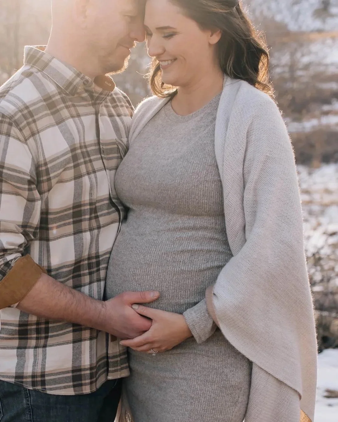 A pregnant woman in a gray dress and a woman in a light-colored cardigan and gray shirt, standing close together outdoors with snow and trees in the background, touching her belly and smiling.