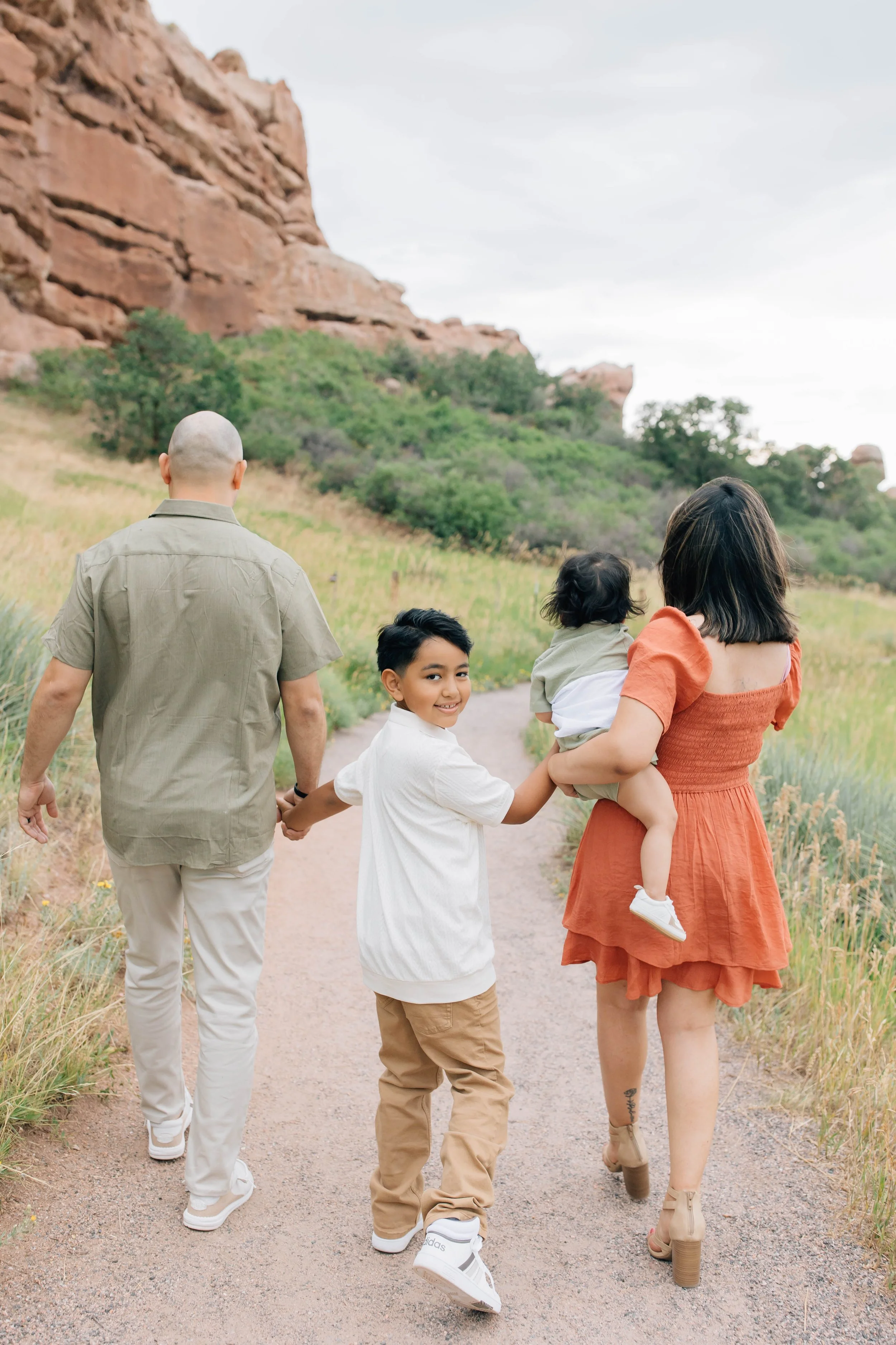 A family of four walking on a dirt trail in a natural, scenic outdoor setting with red rocks and green foliage, hand in hand, with smiling children.