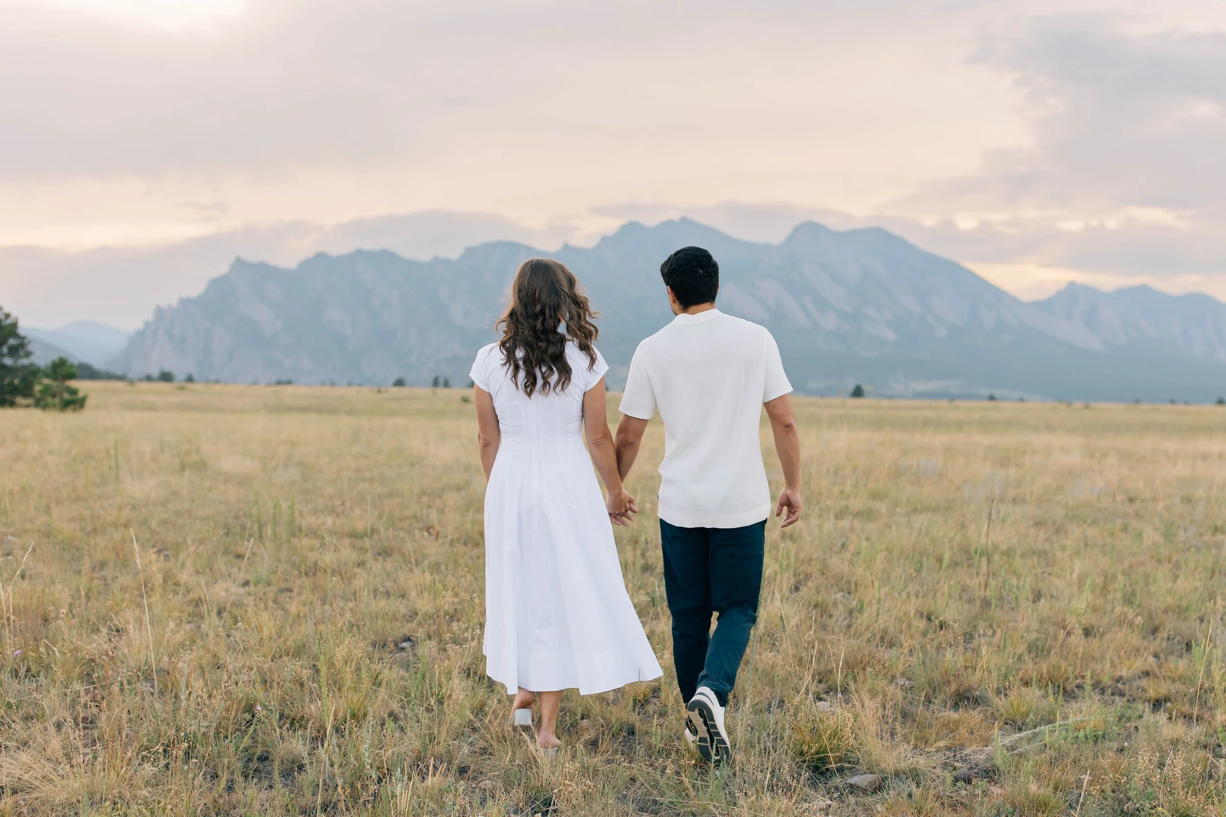 A couple holding hands and walking through a field with mountains in the background.