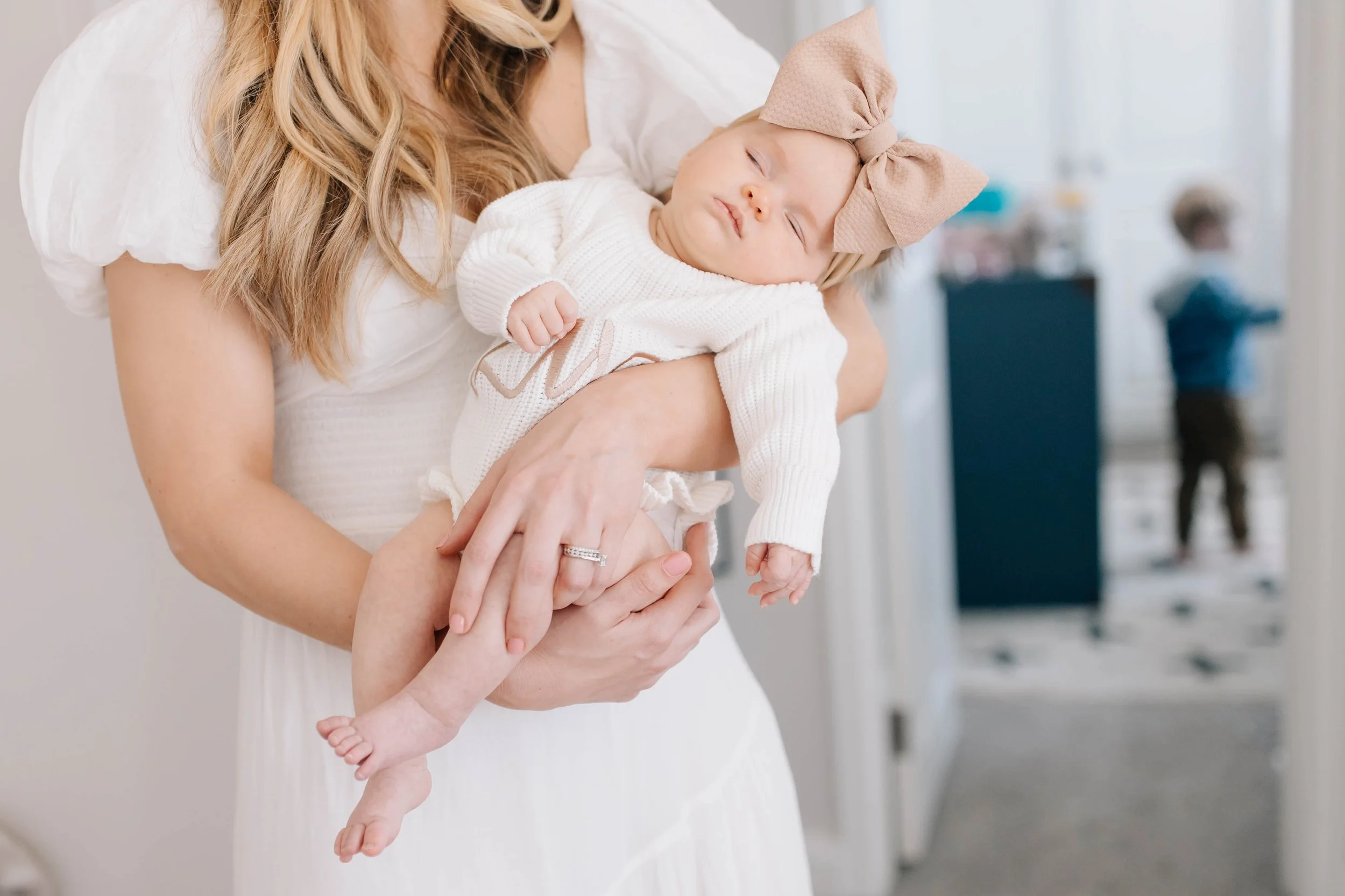 A woman holding a sleeping baby girl with a bow on her head in a white sweater in a home interior.