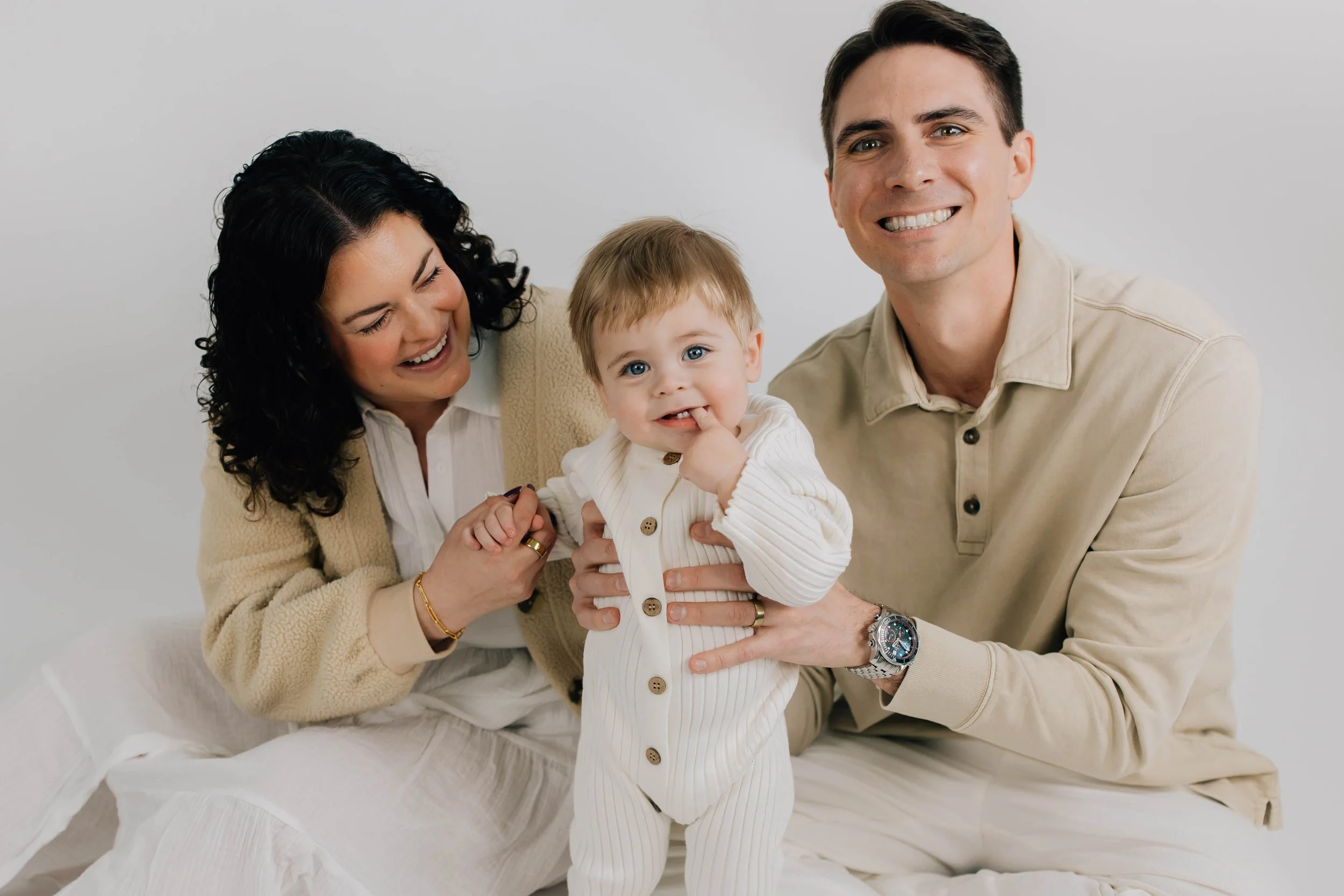 A happy family of three, including a woman, a man, and a toddler, sitting together on a white background, smiling and interacting playfully.