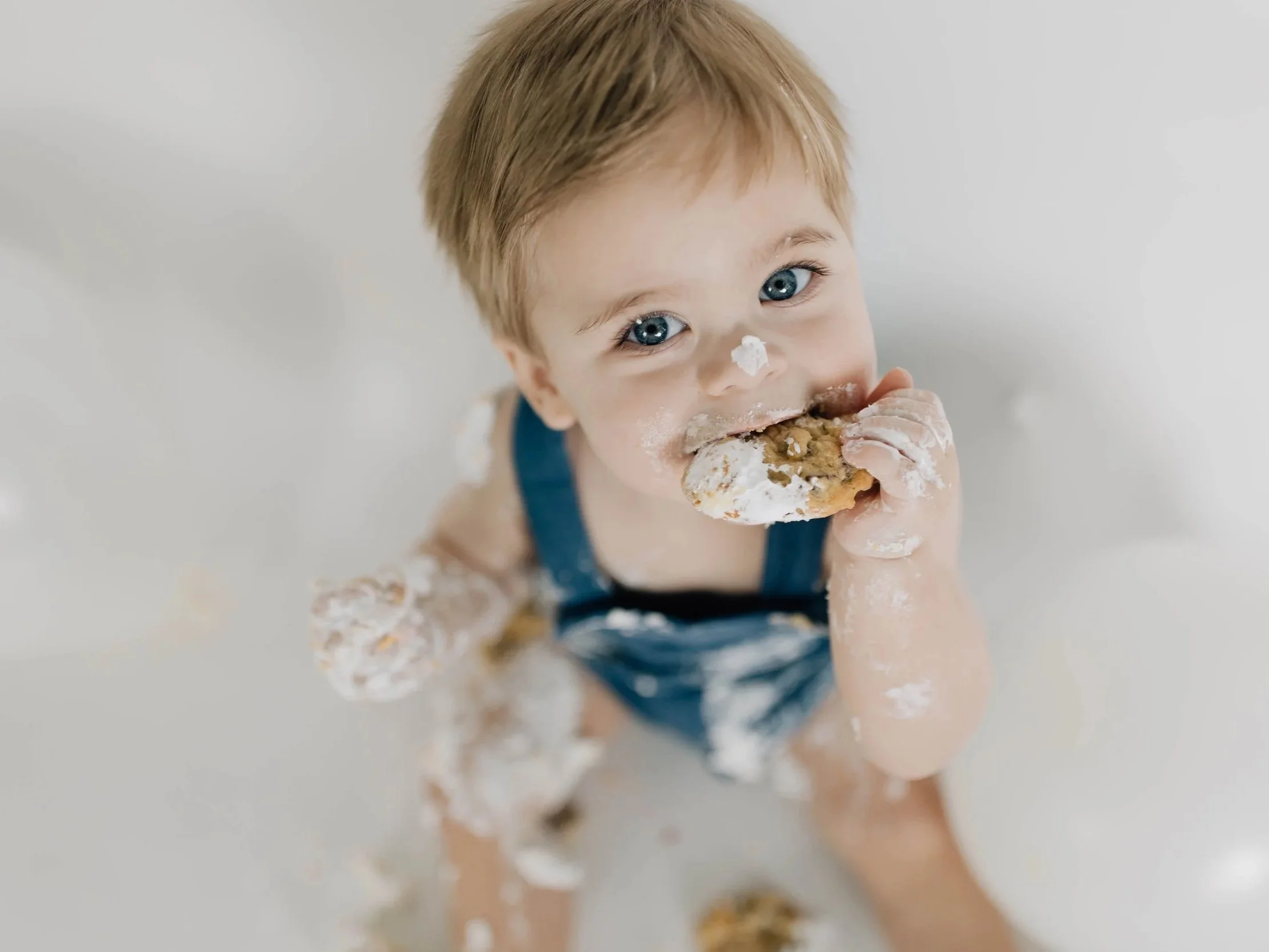 A young child with blue eyes and light brown hair eating a cookie with white frosting, covered in crumbs and frosting, in a bath with white bubbles.