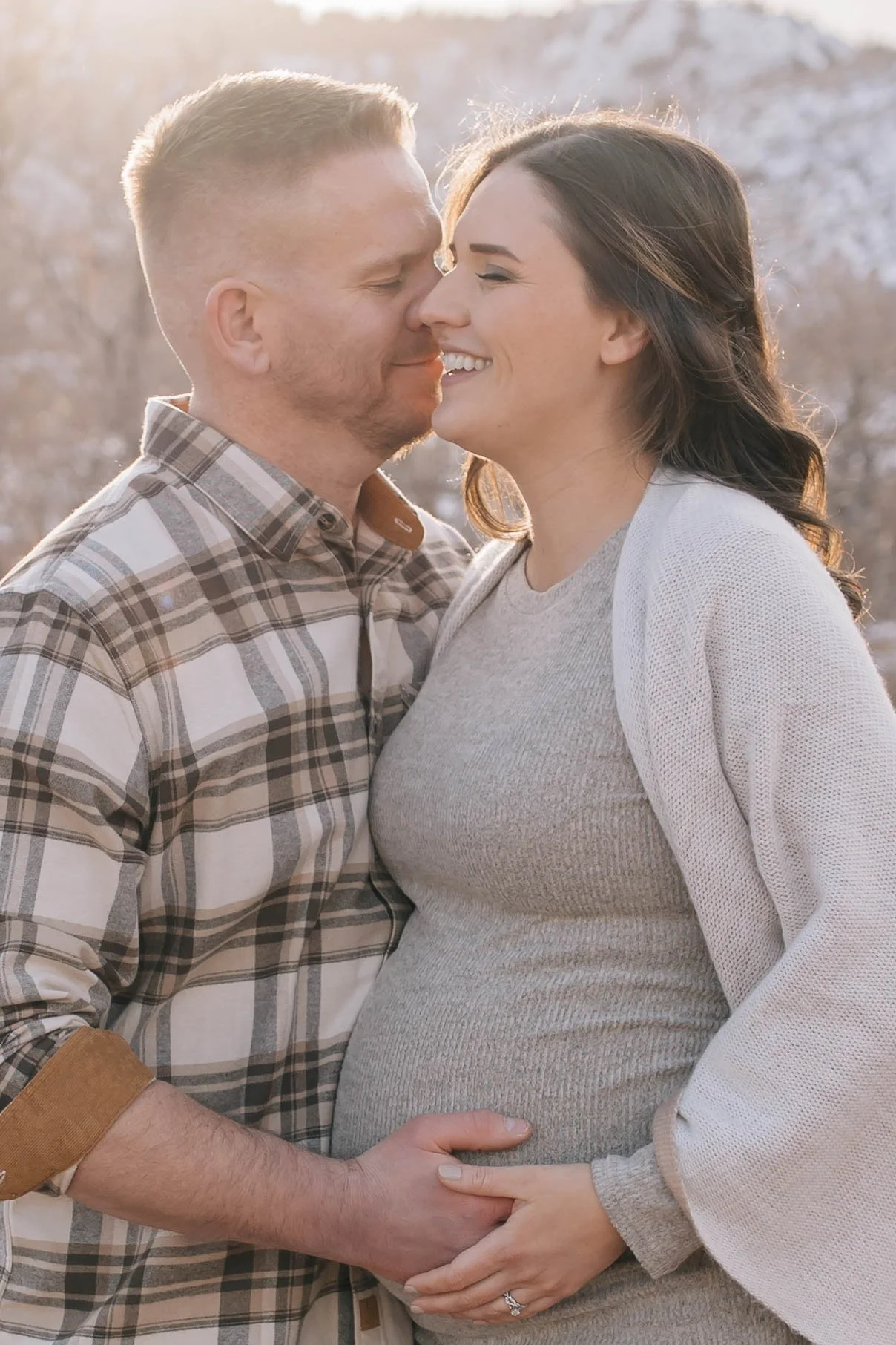 A couple is embracing outdoors, touching foreheads with eyes closed and smiling, with a snowy background and sunlight shining on them.