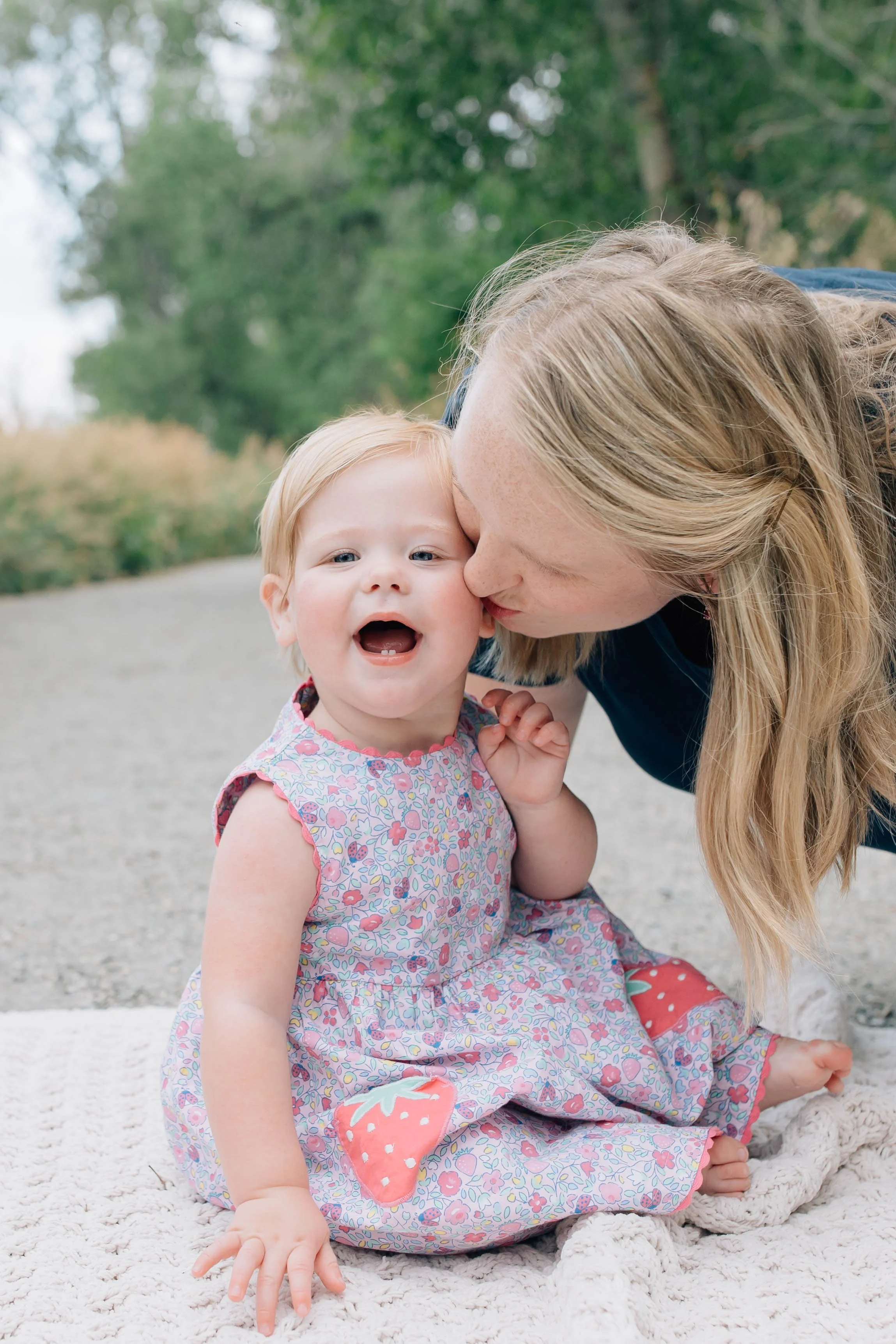 A woman giving a kiss to a young girl, both smiling, outdoors on a blanket with trees in the background.
