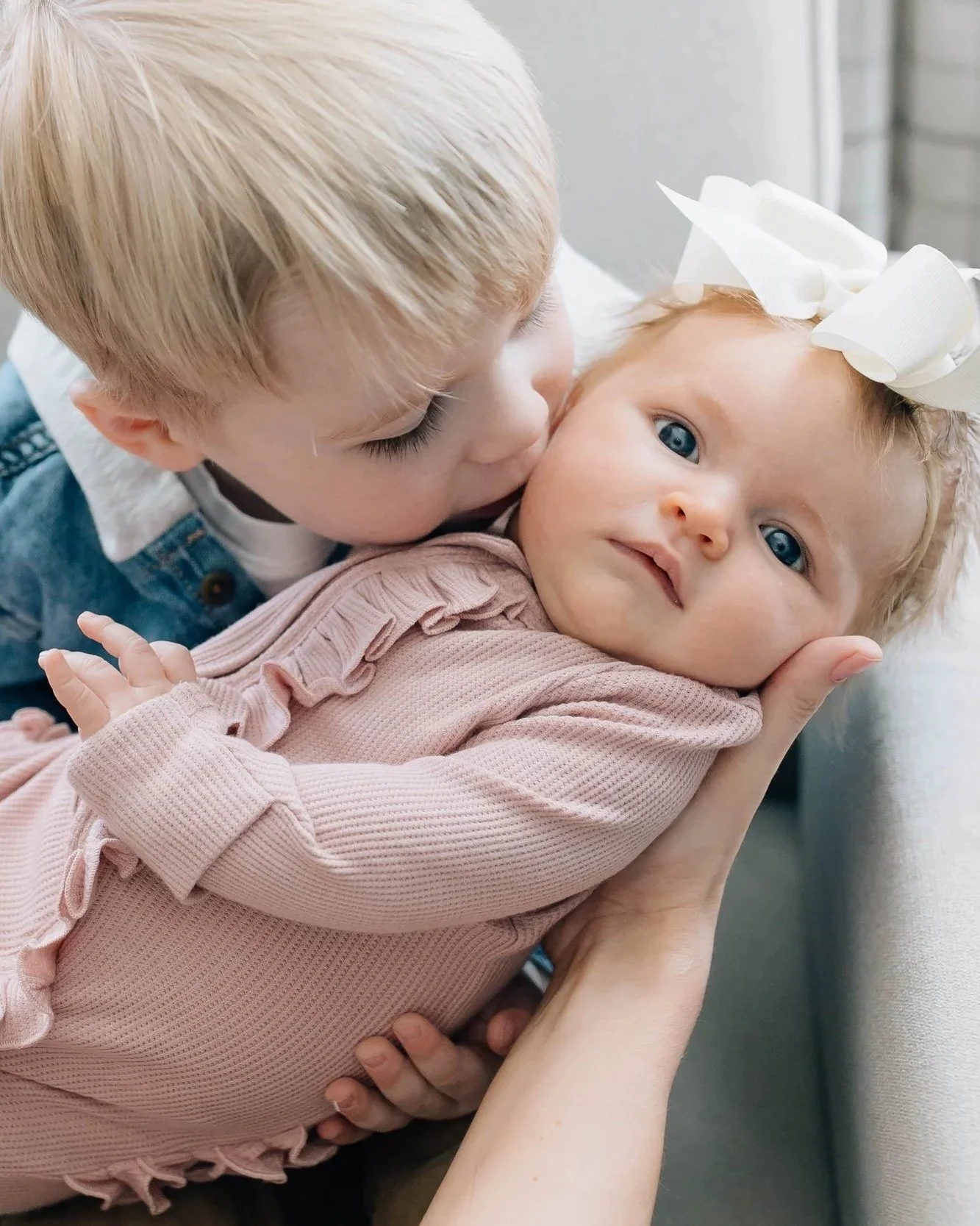 A young boy kissing a baby girl with blue eyes and a white bow in her hair, who is lying on a person's arm.