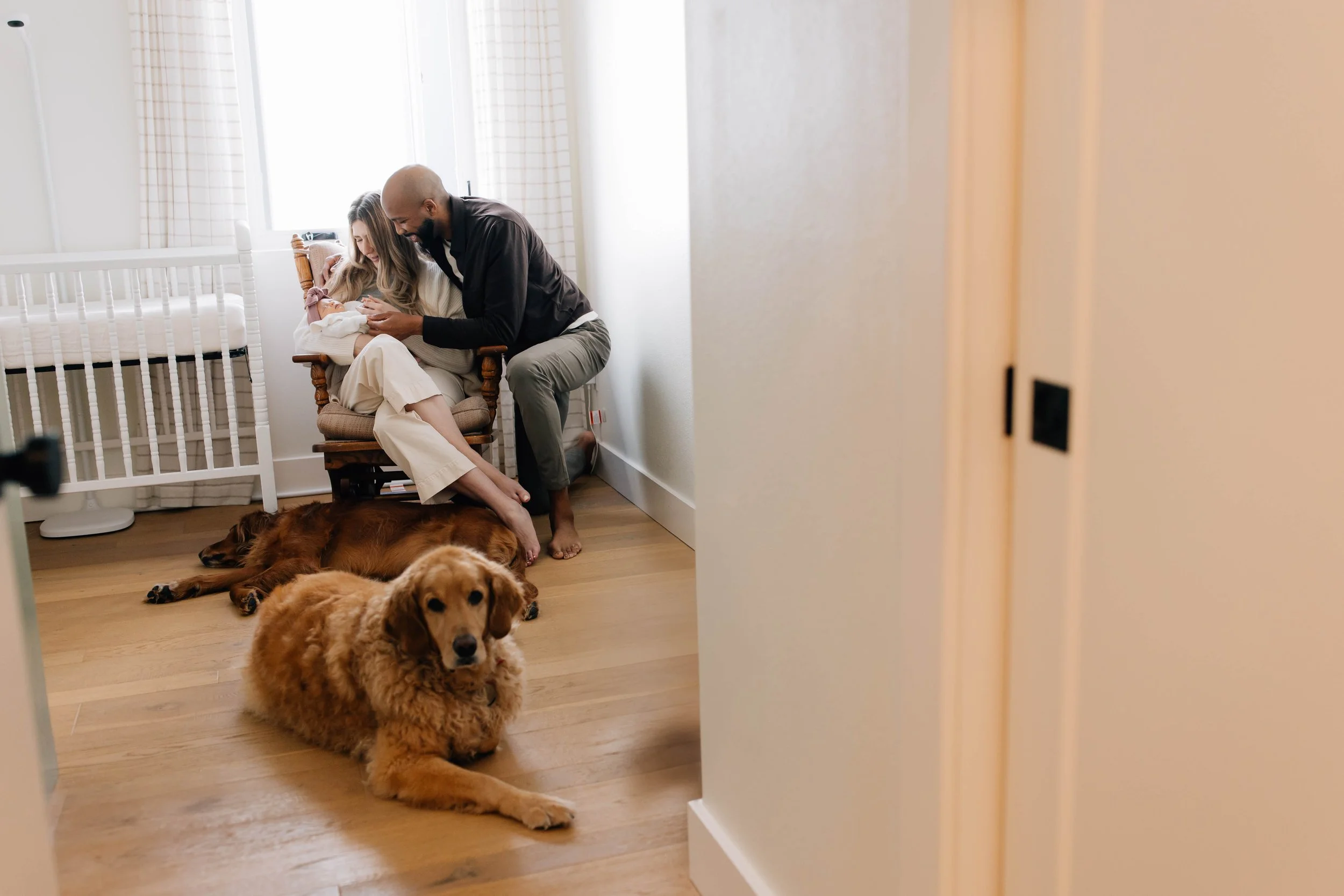 Newborn baby held by a woman sitting in a wooden chair, with a man leaning over, inside a bright room with a crib and two dogs on the floor.